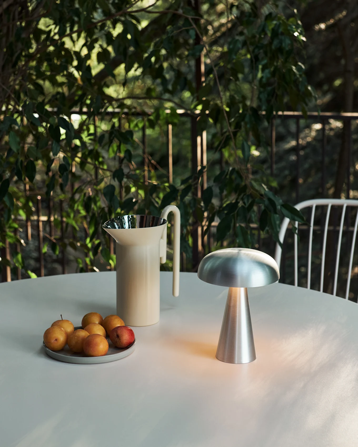 An outdoor table setting with a glowing silver mushroom lamp, a cream pitcher, and a plate of plums amidst green foliage.