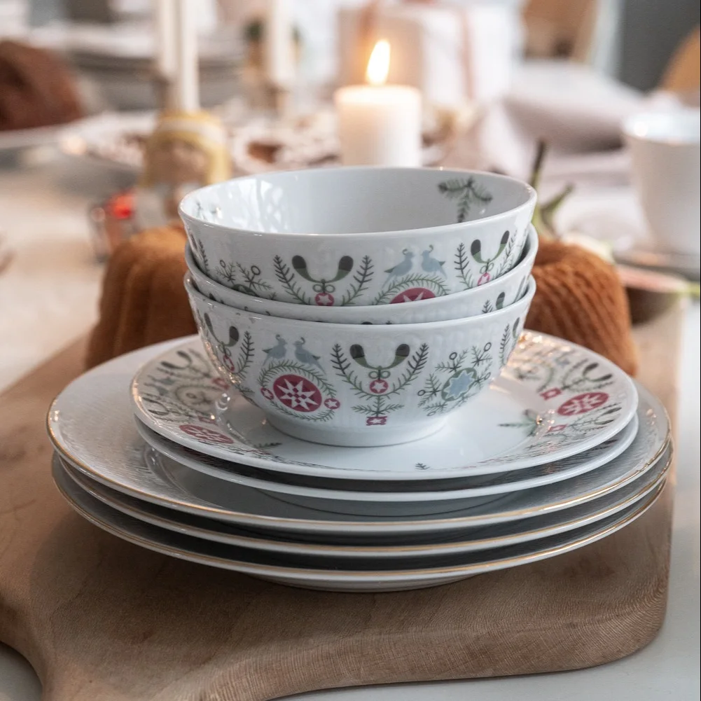 Stack of white ceramic bowls and plates with festive folk-art patterns on a wooden board.