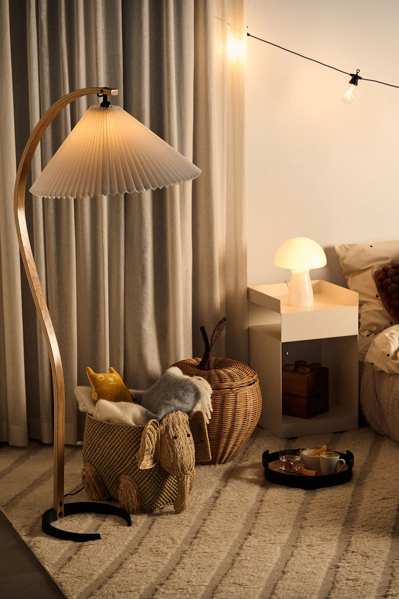 A curved floor lamp stands in a children's room along with a mushroom lamp and a set of string lights. 