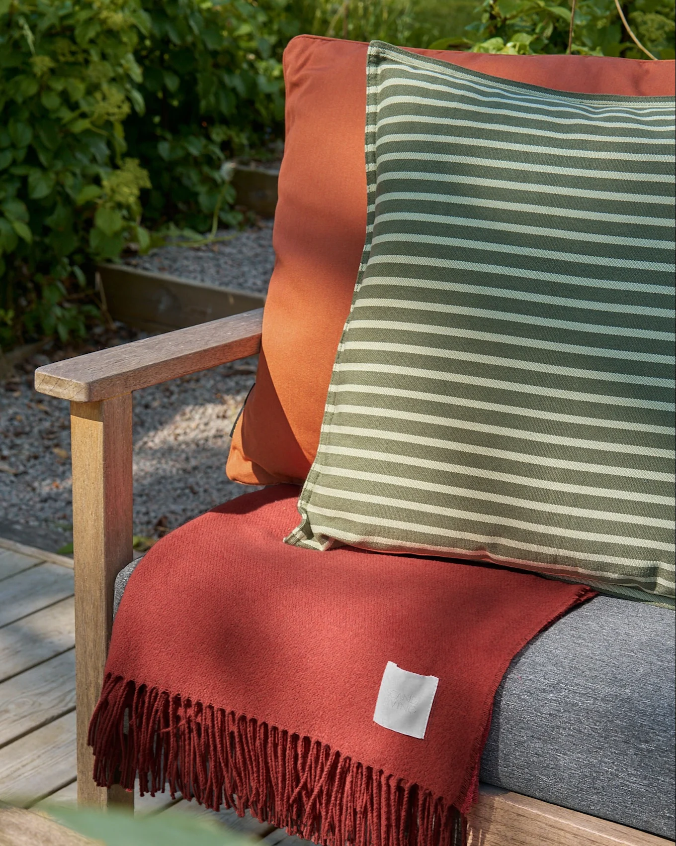 Outdoor wooden chair with an orange cushion, a striped green and white pillow, and a terracotta fringed blanket.