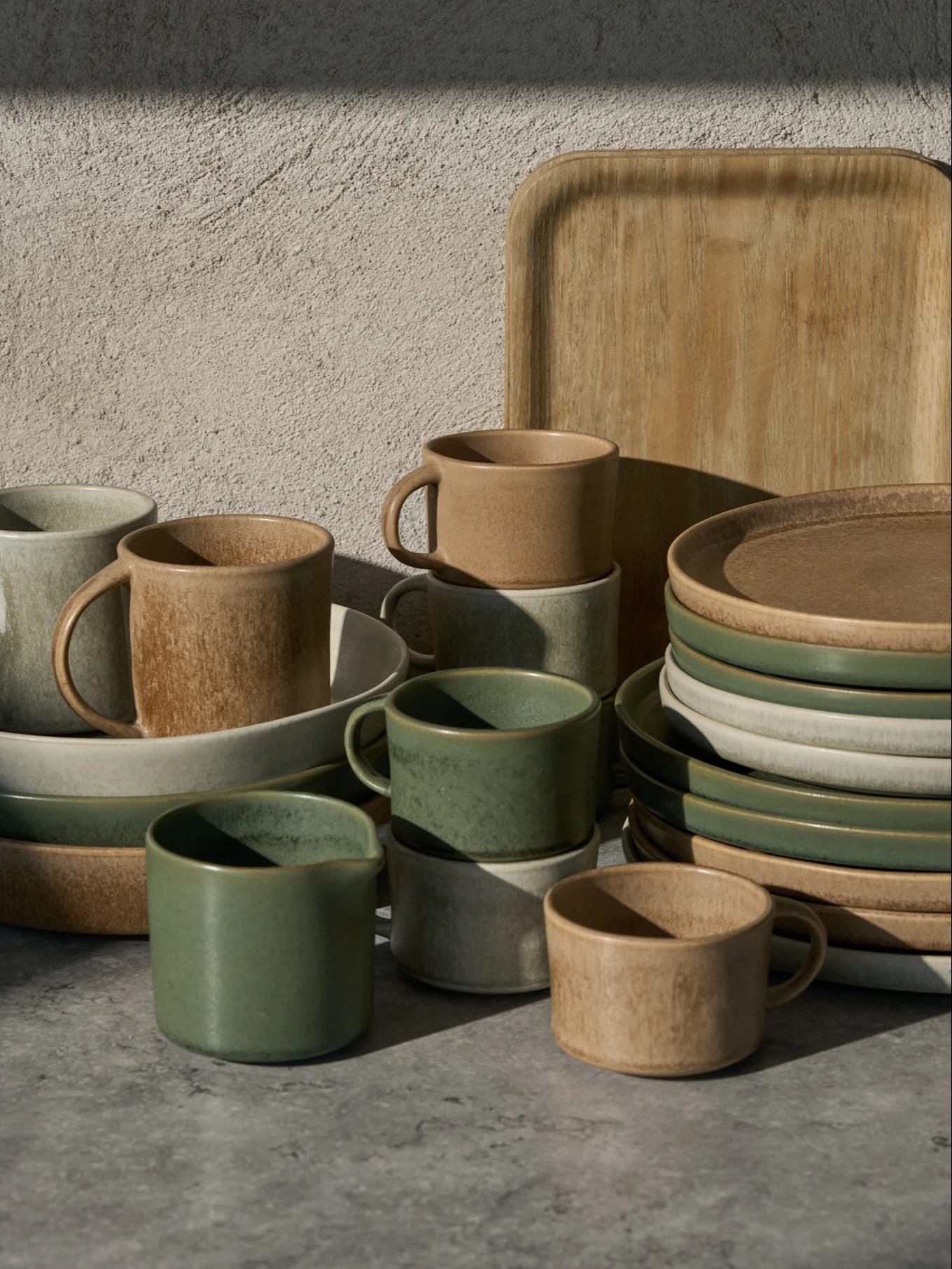 Collection of green, beige, and light gray ceramic cups, bowls, and plates, with a wooden tray, on a gray surface.