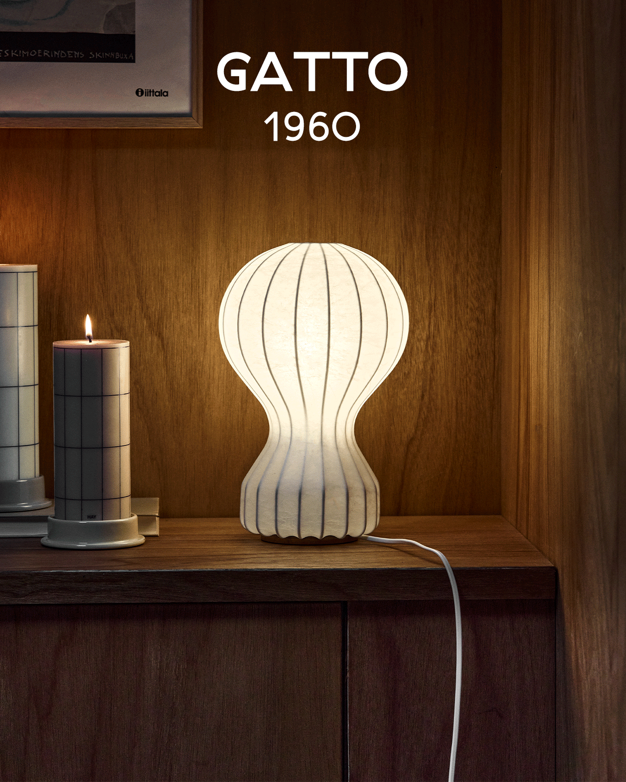 A glowing white ribbed lamp with an hourglass shape sits on a wooden shelf next to a lit candle. Text: GATTO 1960.