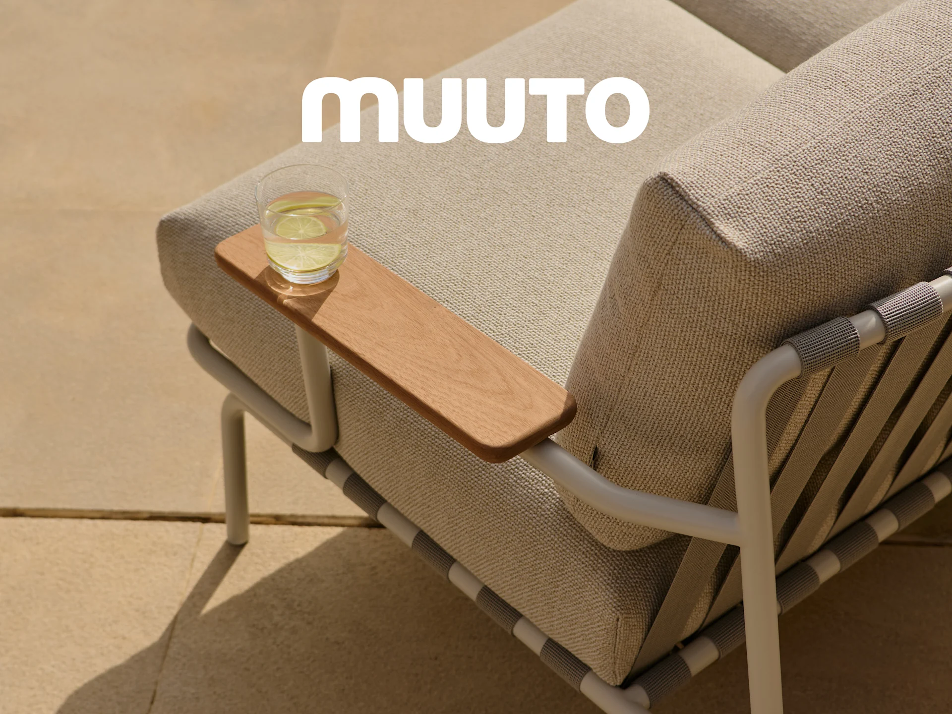 Close-up of an outdoor sofa with light fabric cushions, a wooden armrest with a glass of water, and the white Muuto logo.