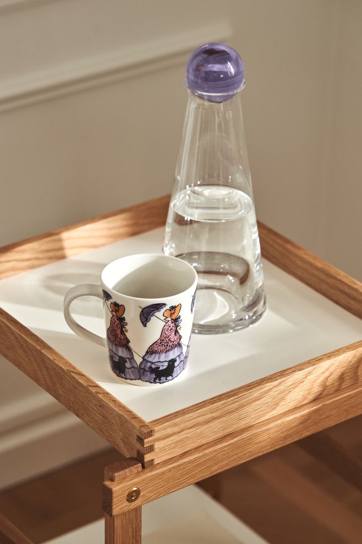 Mug with the Elsa Beskow motif Aunt Gredelin from Design House Stockholm and Fia carafe in glass with purple lid on Frame shelf in oak and white.