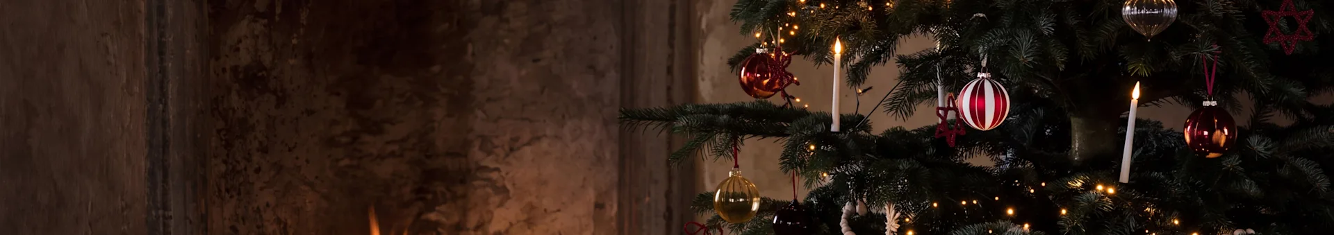 A festive Christmas tree with ornaments and a lit candle next to a warm fireplace with burning logs.