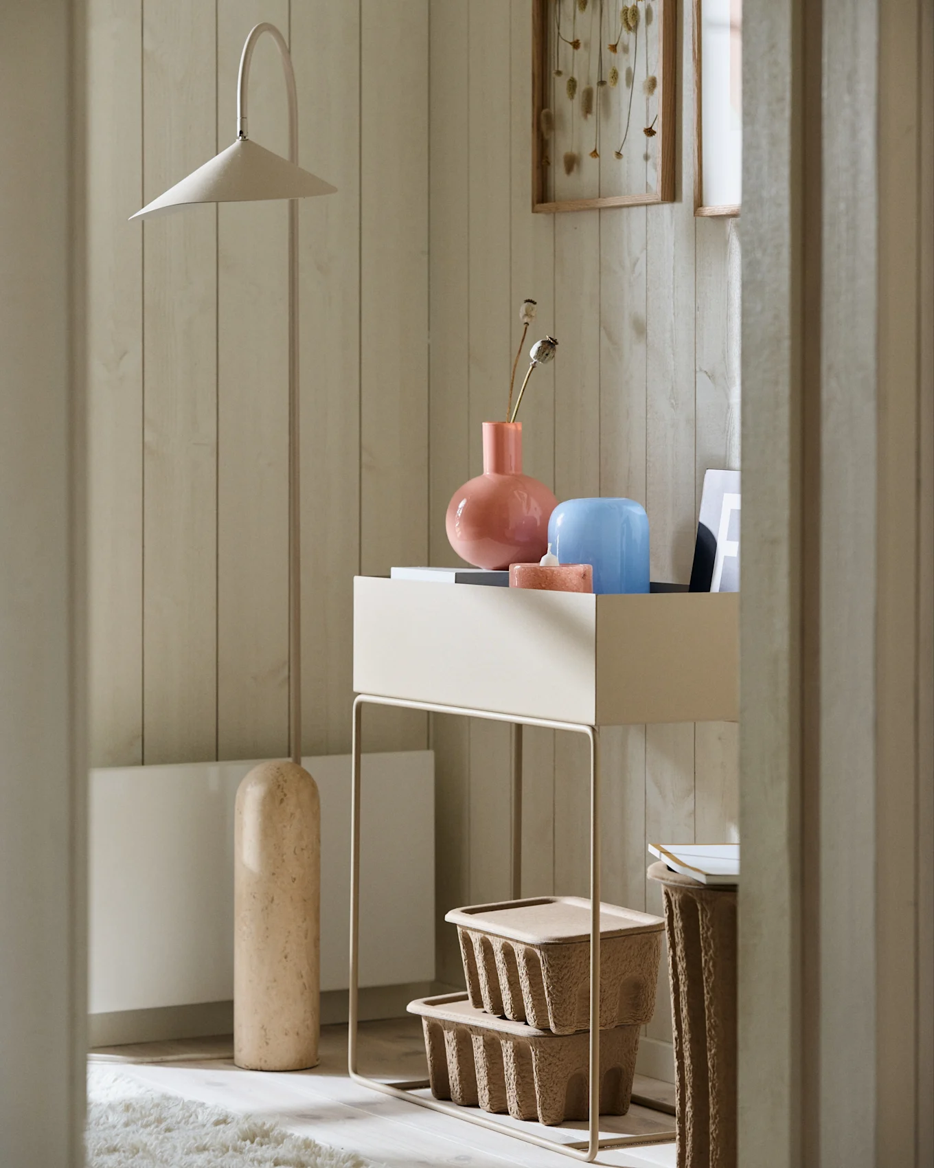 Minimalist corner with a tall beige floor lamp, a console table featuring pink and blue vases, and textured brown storage boxes.