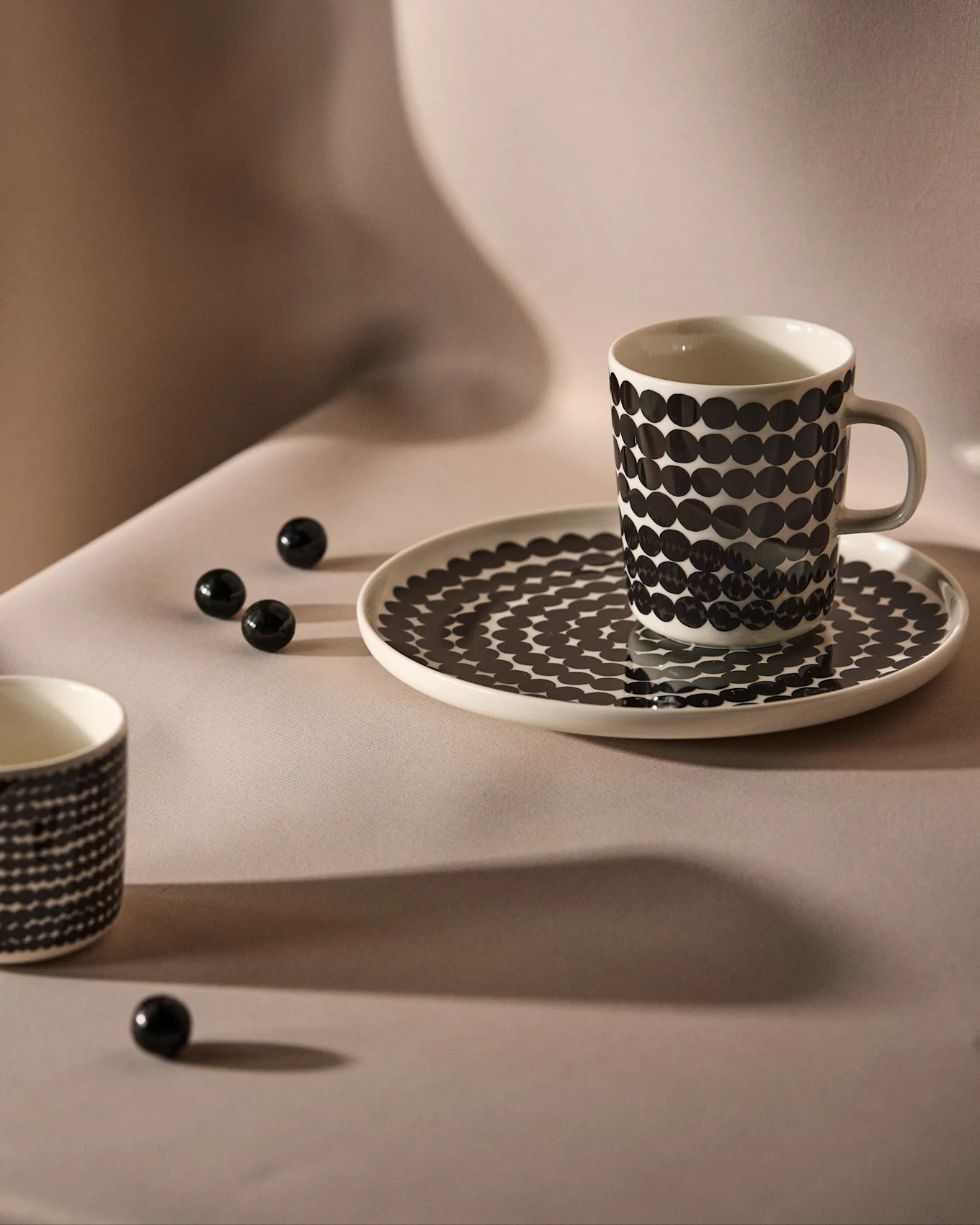 White mug and saucer with black polka dot pattern on a neutral background, with scattered black decorative beads.