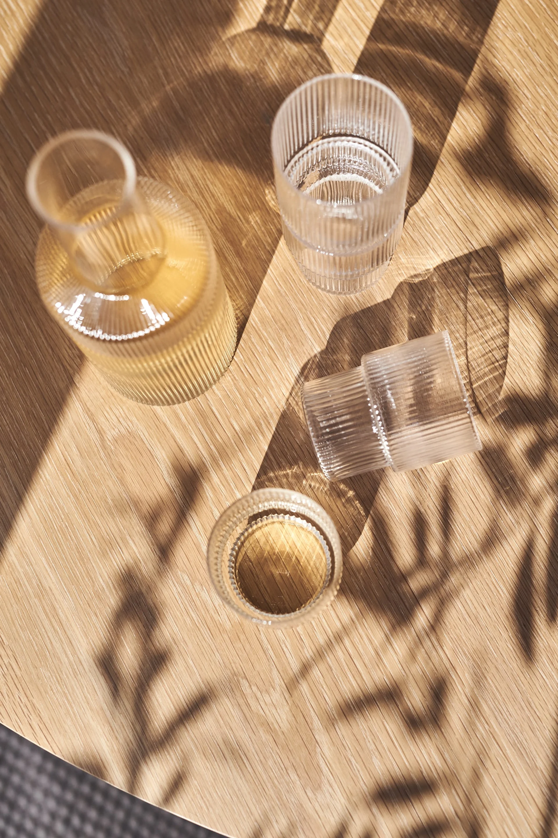 A ribbed glass bottle and two ribbed glasses, one with liquid, on a sunlit wooden table with leaf shadows.