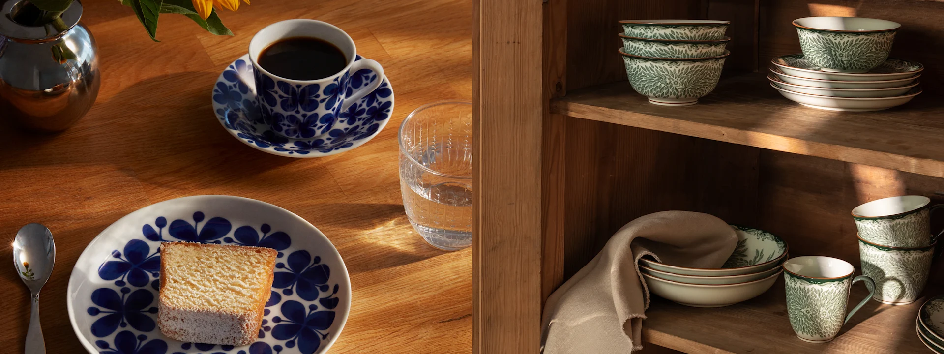 A table setting with a patterned coffee cup, cake, and water, next to a wooden cabinet filled with green patterned dinnerware.