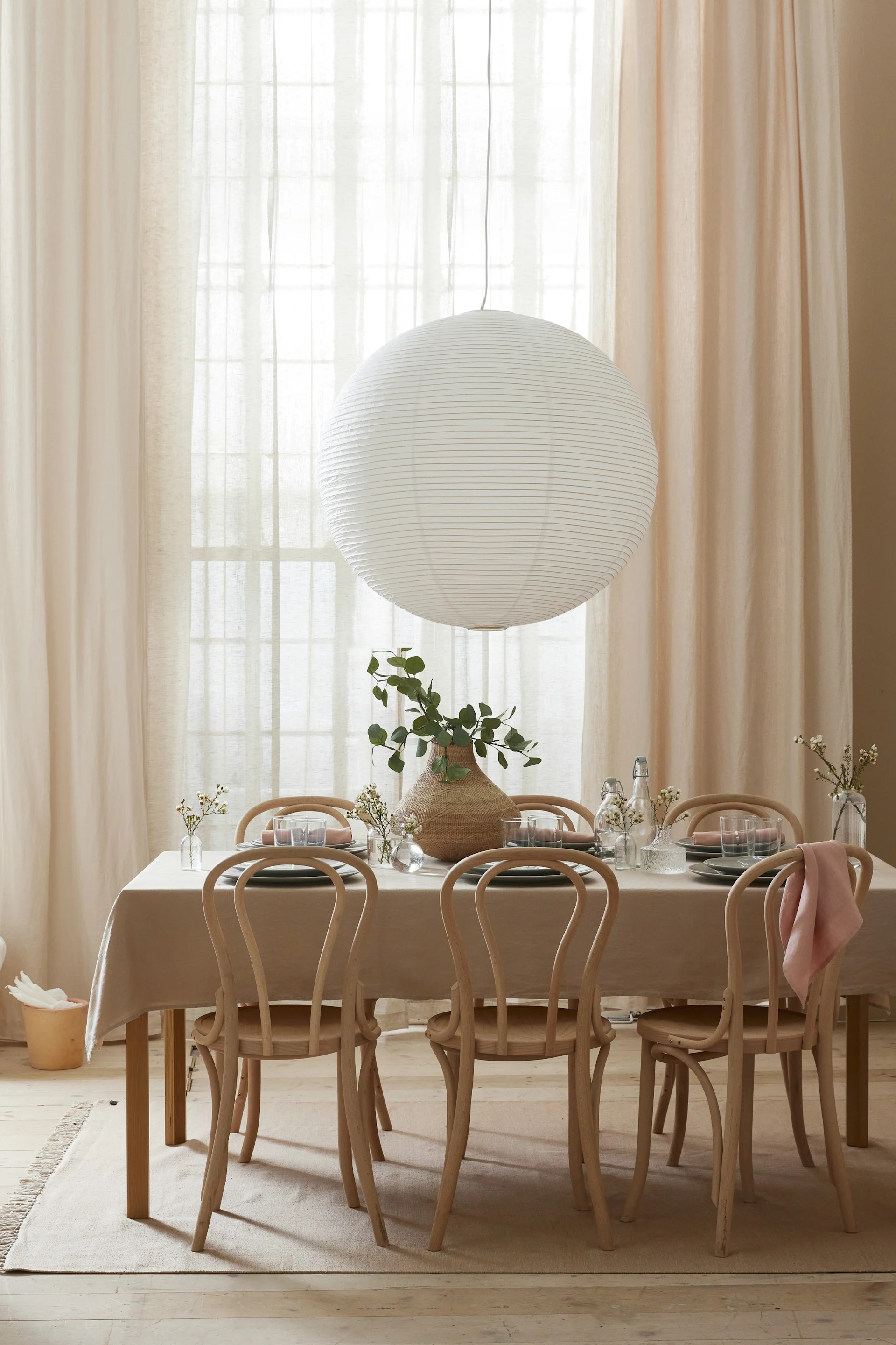 Dining room with a table set for six, featuring a large white paper lantern, wooden chairs, and a centerpiece with green branches.