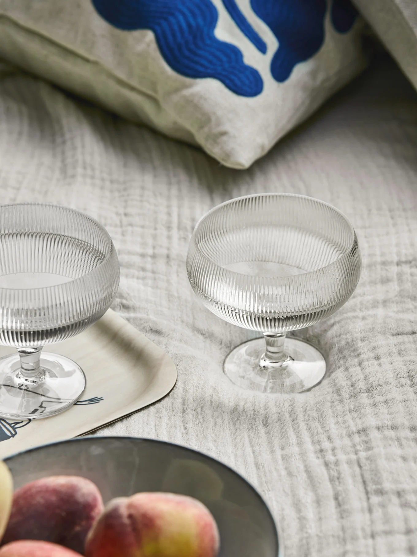 Two ribbed glass dessert bowls, a pillow with blue abstract art, a wooden tray, and peaches on a light fabric.
