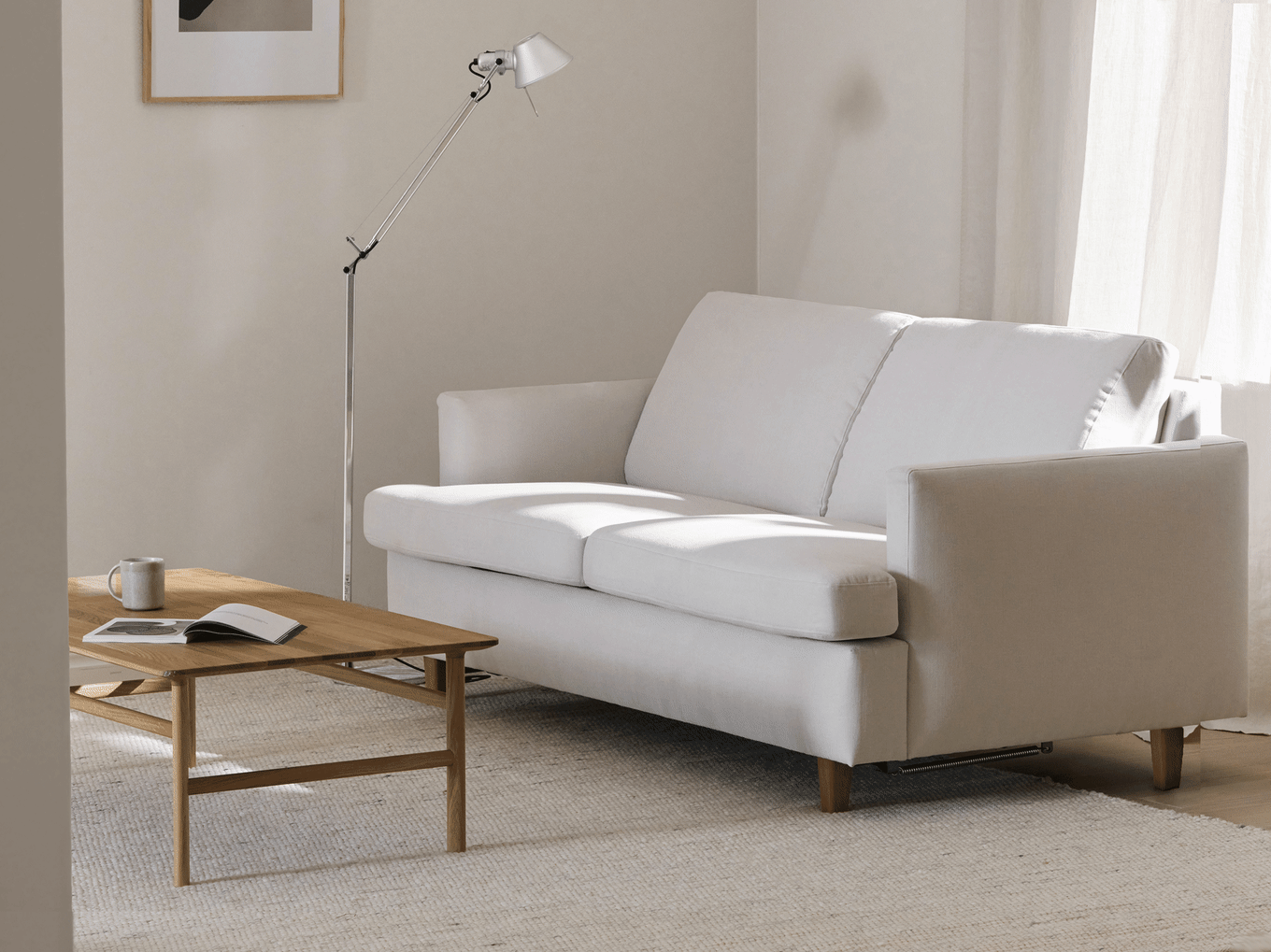 A minimalist living room with a light grey two-seater sofa, a wooden coffee table, a modern floor lamp, and sheer curtains.