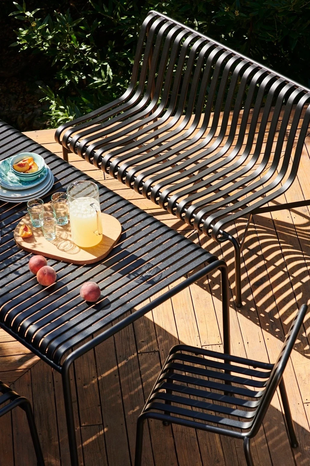 Black slatted outdoor furniture set on a wooden deck with a bench, table, and chair, featuring lemonade and peaches.