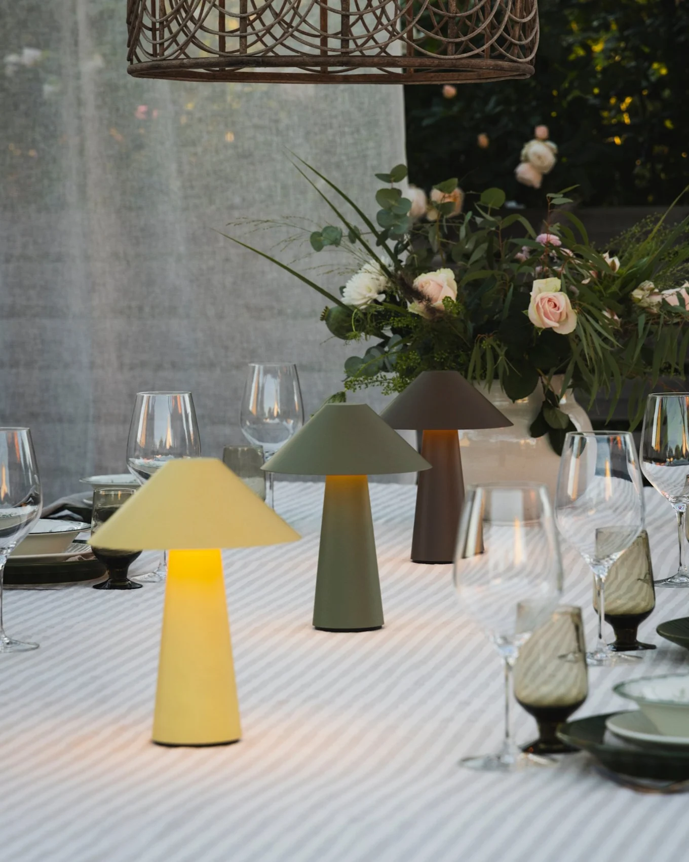 Outdoor dining table with three illuminated mushroom lamps, floral centerpiece, and a wicker pendant light.