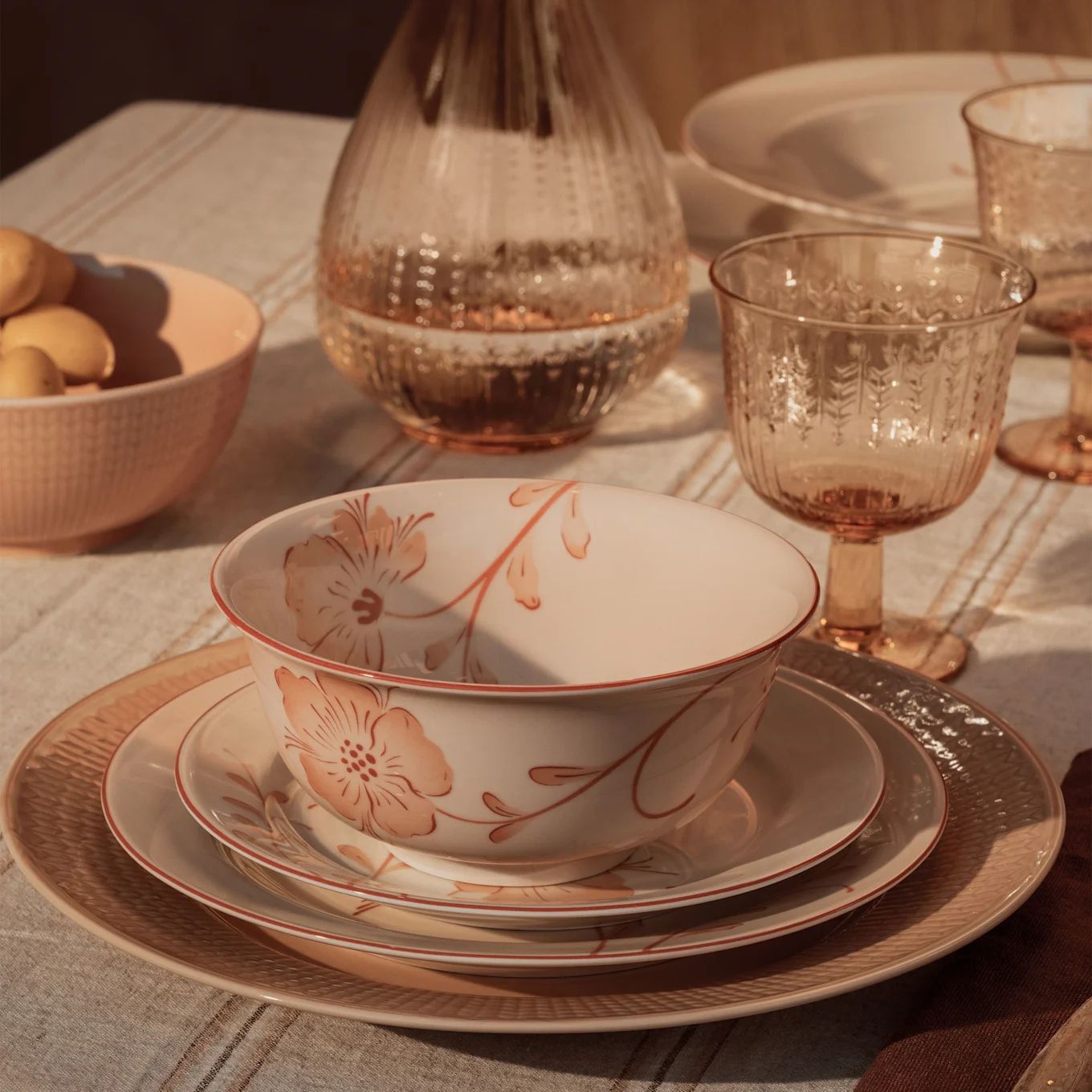 A rustic table setting with a floral bowl, stacked plates, amber glasses, and a peach bowl of produce on a striped tablecloth.