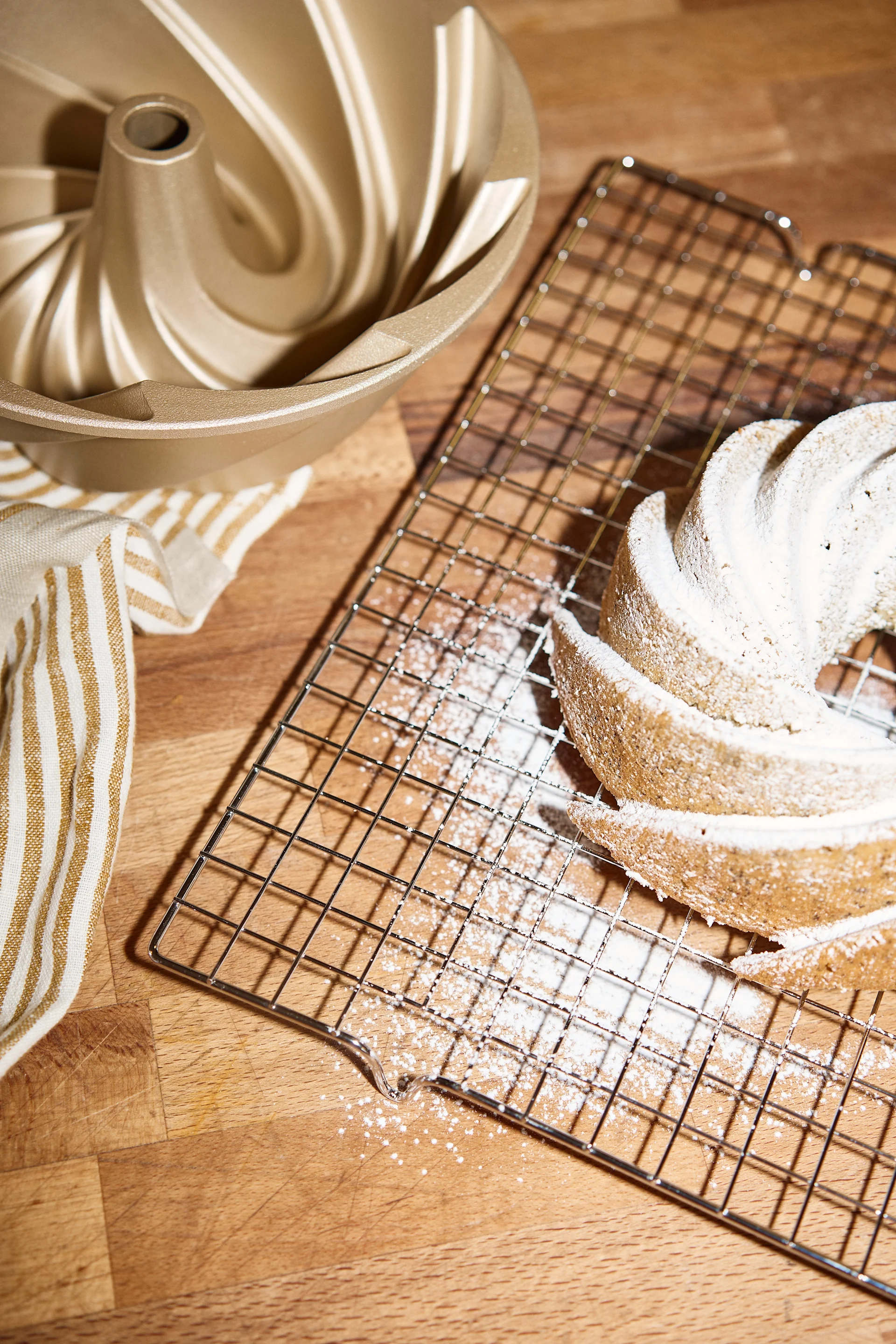 A golden bundt pan, a striped towel, and a bundt cake dusted with powdered sugar on a cooling rack.