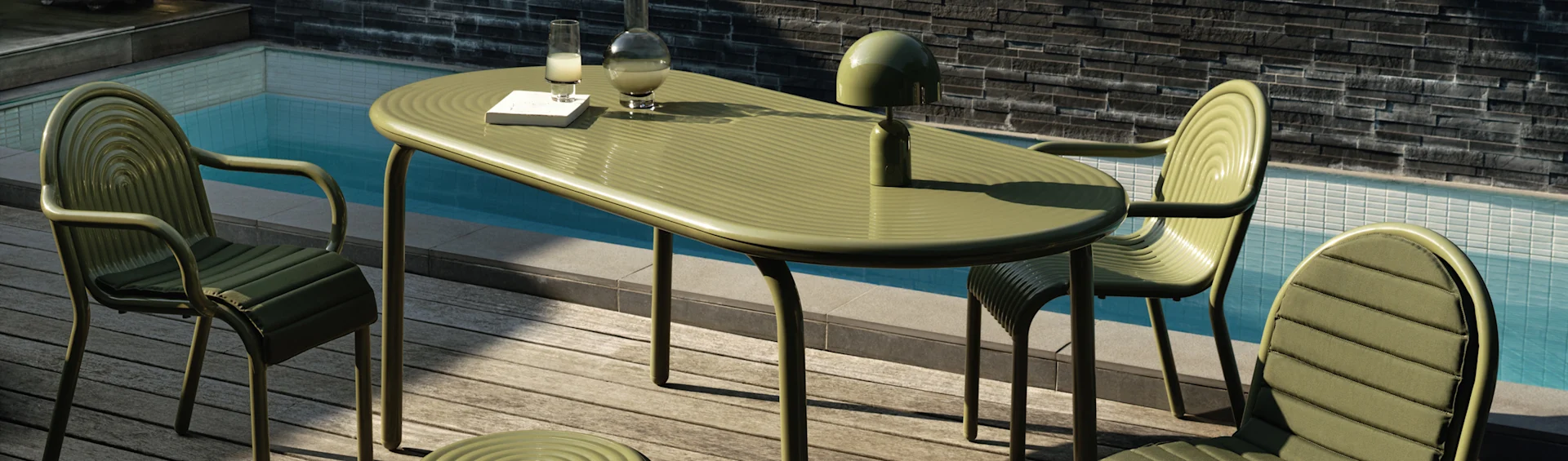 Green ribbed outdoor dining table and chairs on a wooden deck by a pool. A lamp, carafe, glass, and book are on the table.