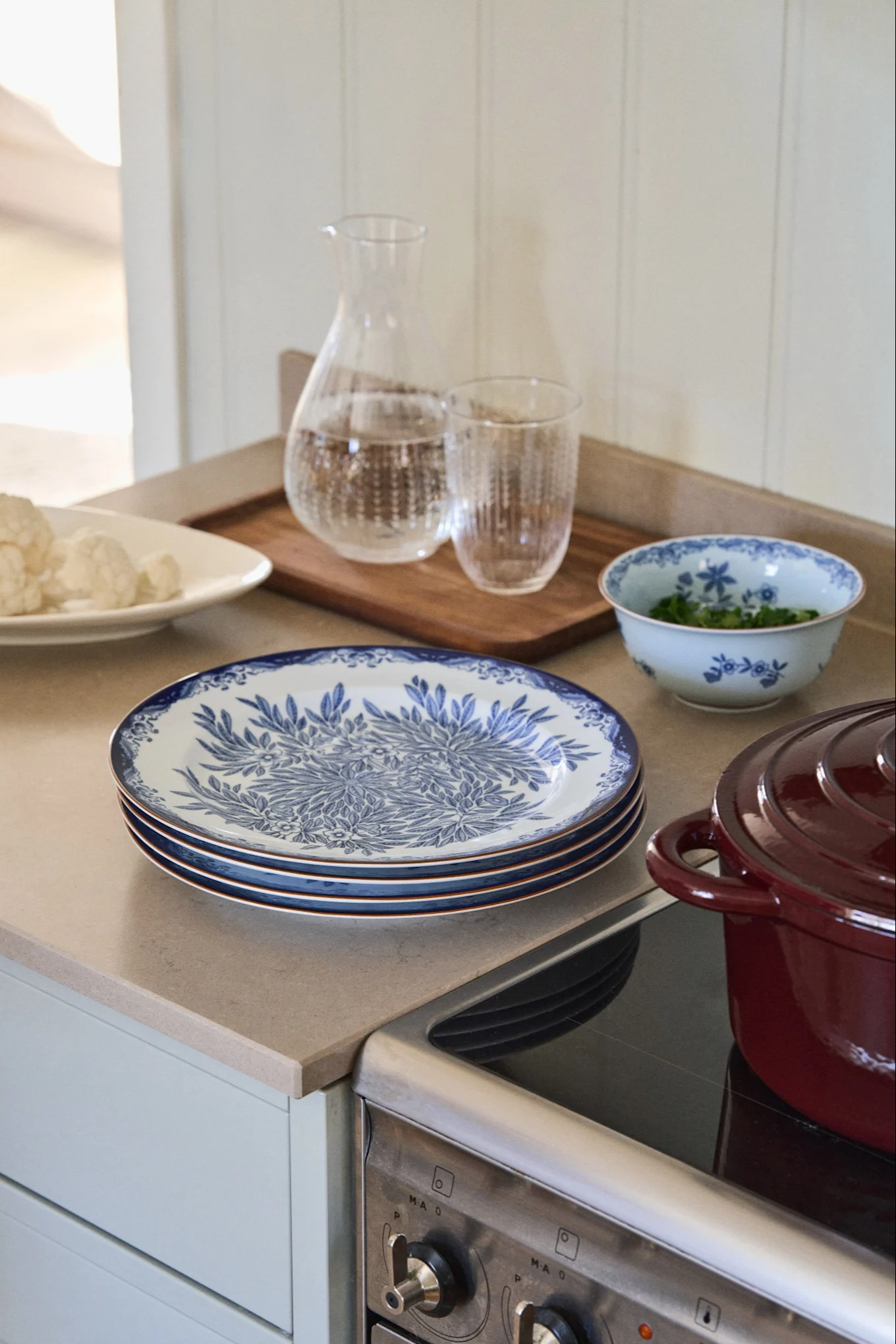 Stack of blue and white patterned plates, a water carafe, and a red pot on a kitchen counter with cauliflower and greens.