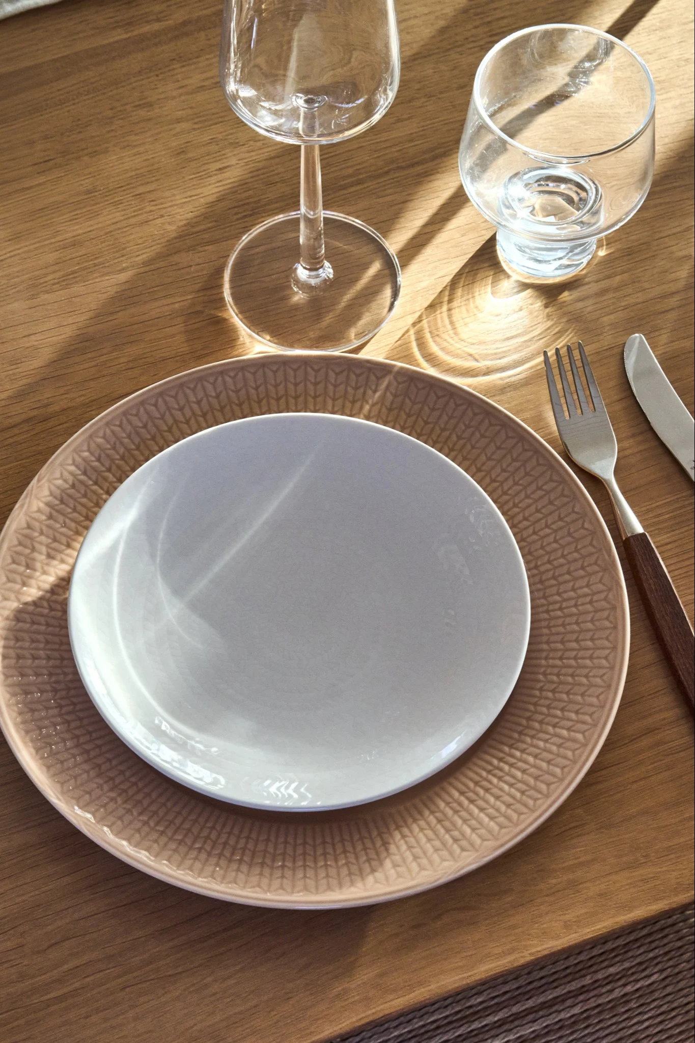 A table setting with a light plate on a patterned beige plate, a fork, a knife with a wooden handle, and two glasses on a wooden table.