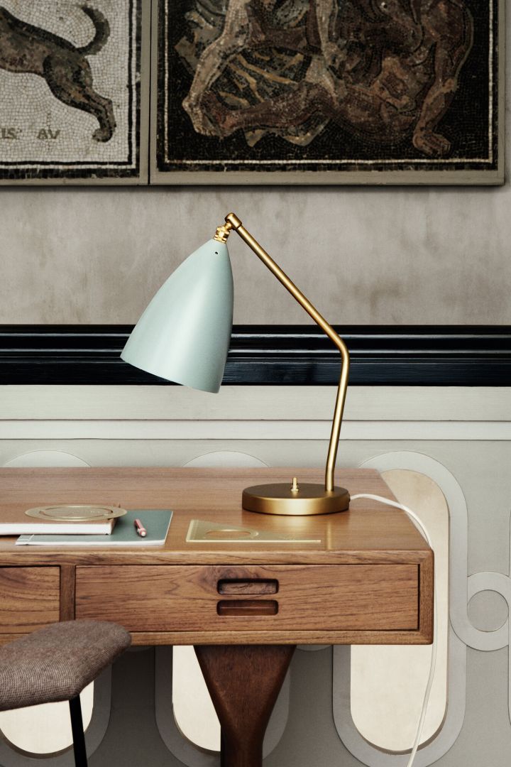 Here you see the Grasshopper table lamp in brass with a blue-grey shade sitting on a wooden desk. 