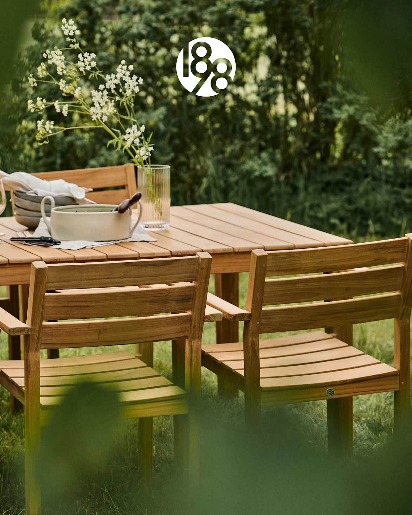 Wooden outdoor dining set with a table and chairs in a lush green garden, featuring white flowers and serving dishes.