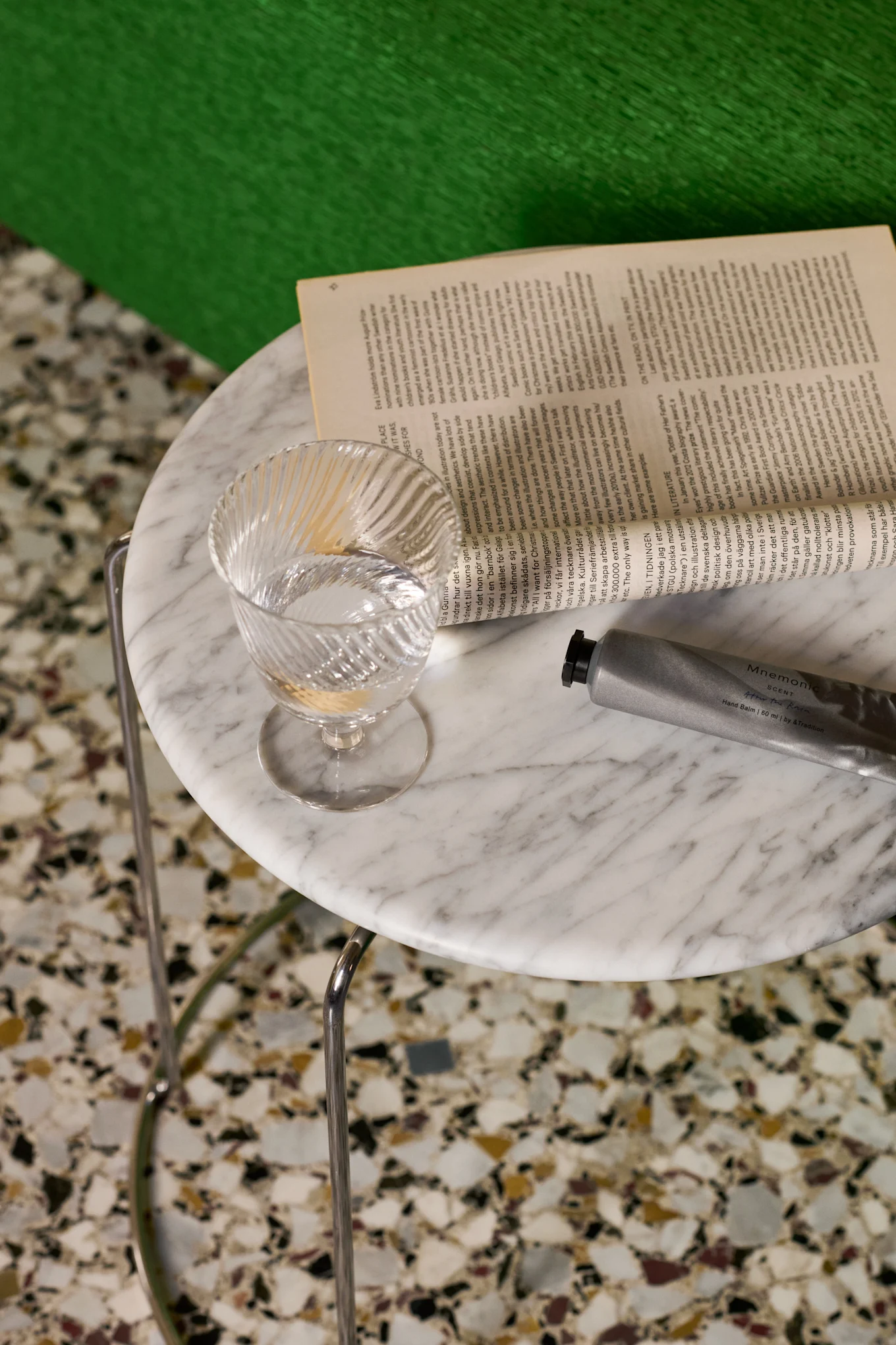 A clear, ribbed glass of water, an open book, and a silver hand balm tube on a marble table.