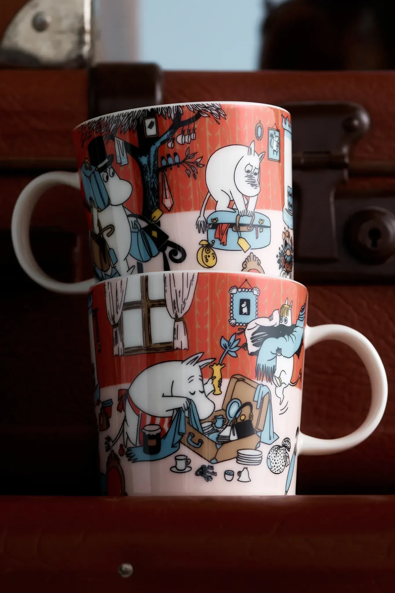 Two stacked red Moomin mugs with illustrations of characters packing suitcases and hanging items on a tree.