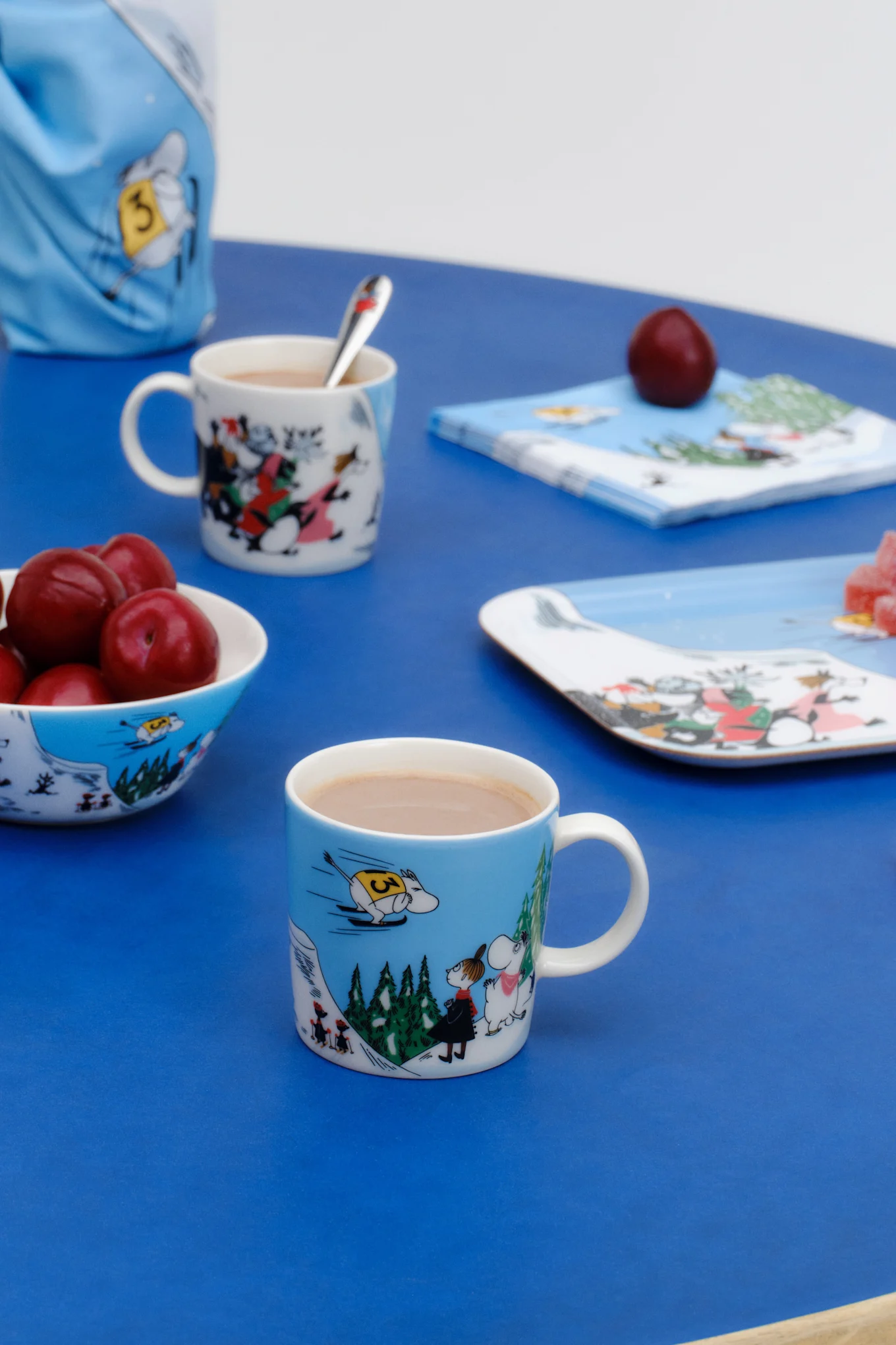 The Moomin mug Winter 2024, ‘Ski Jumping’, stands on a table set in blue.