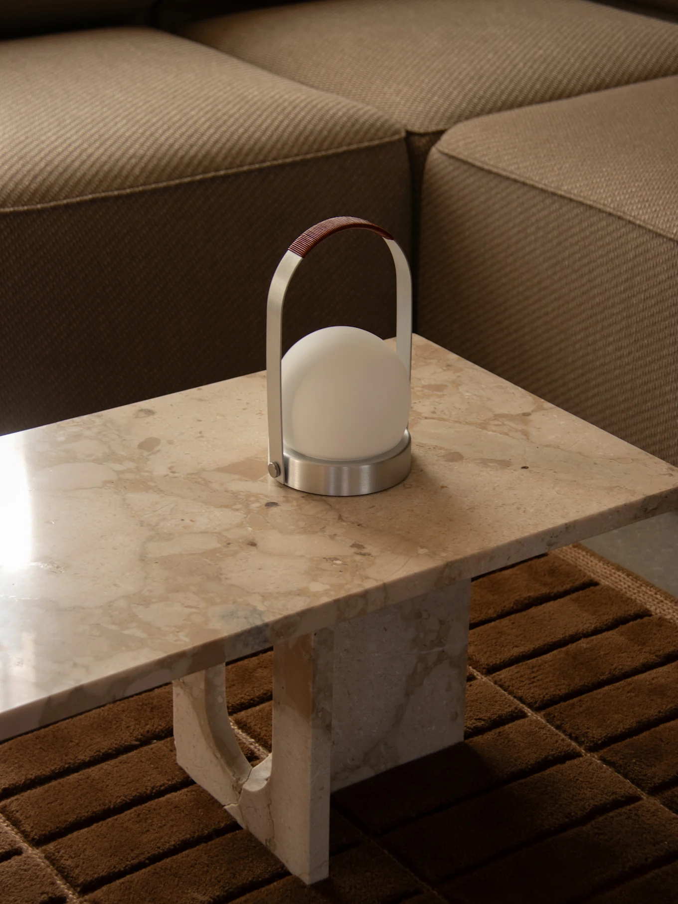 Modern portable lamp with a spherical white light, silver frame, and brown leather handle on a marble table next to a sofa.