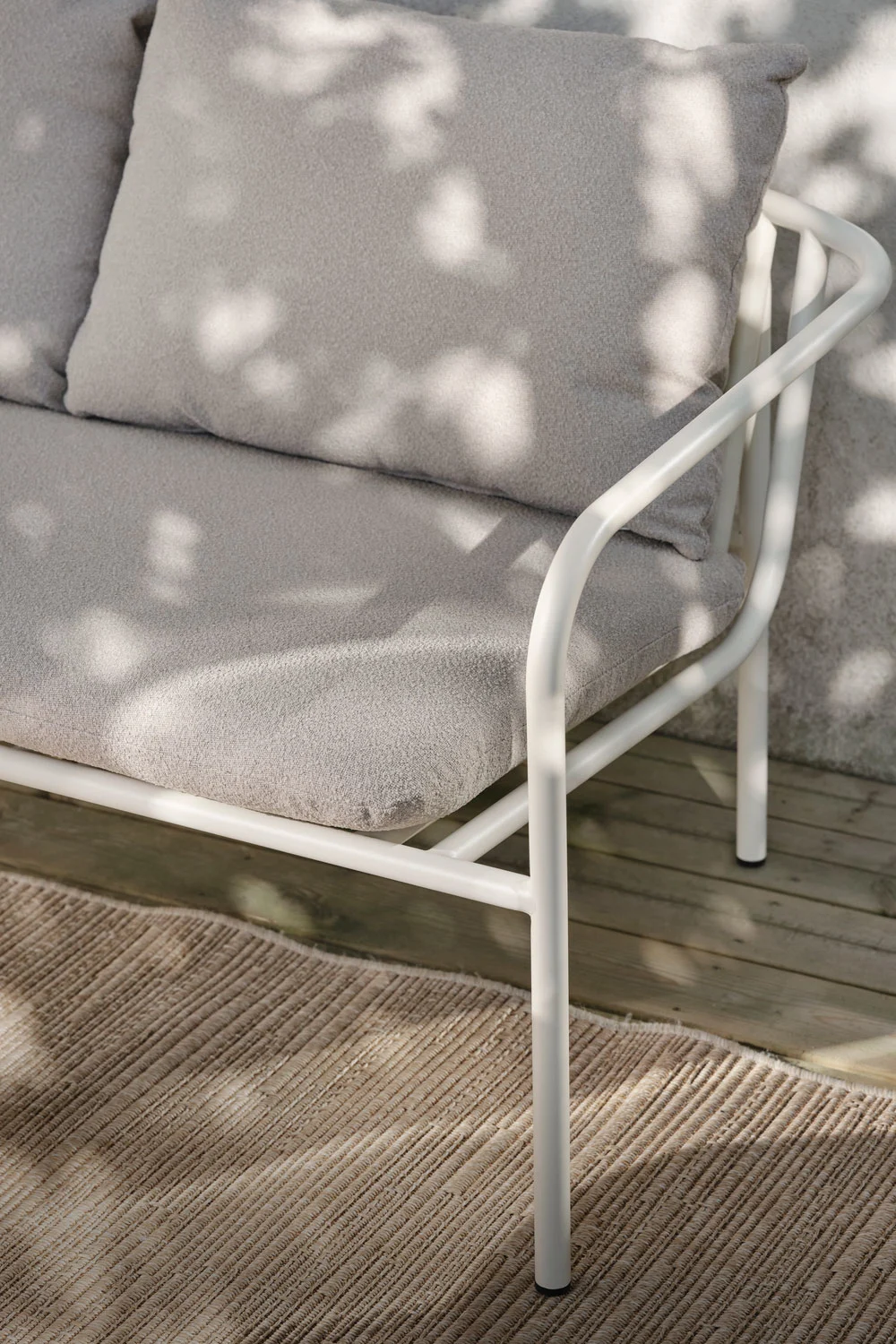Close-up of a white outdoor sofa with light gray cushions, on a wooden deck with a woven rug, dappled in sunlight.