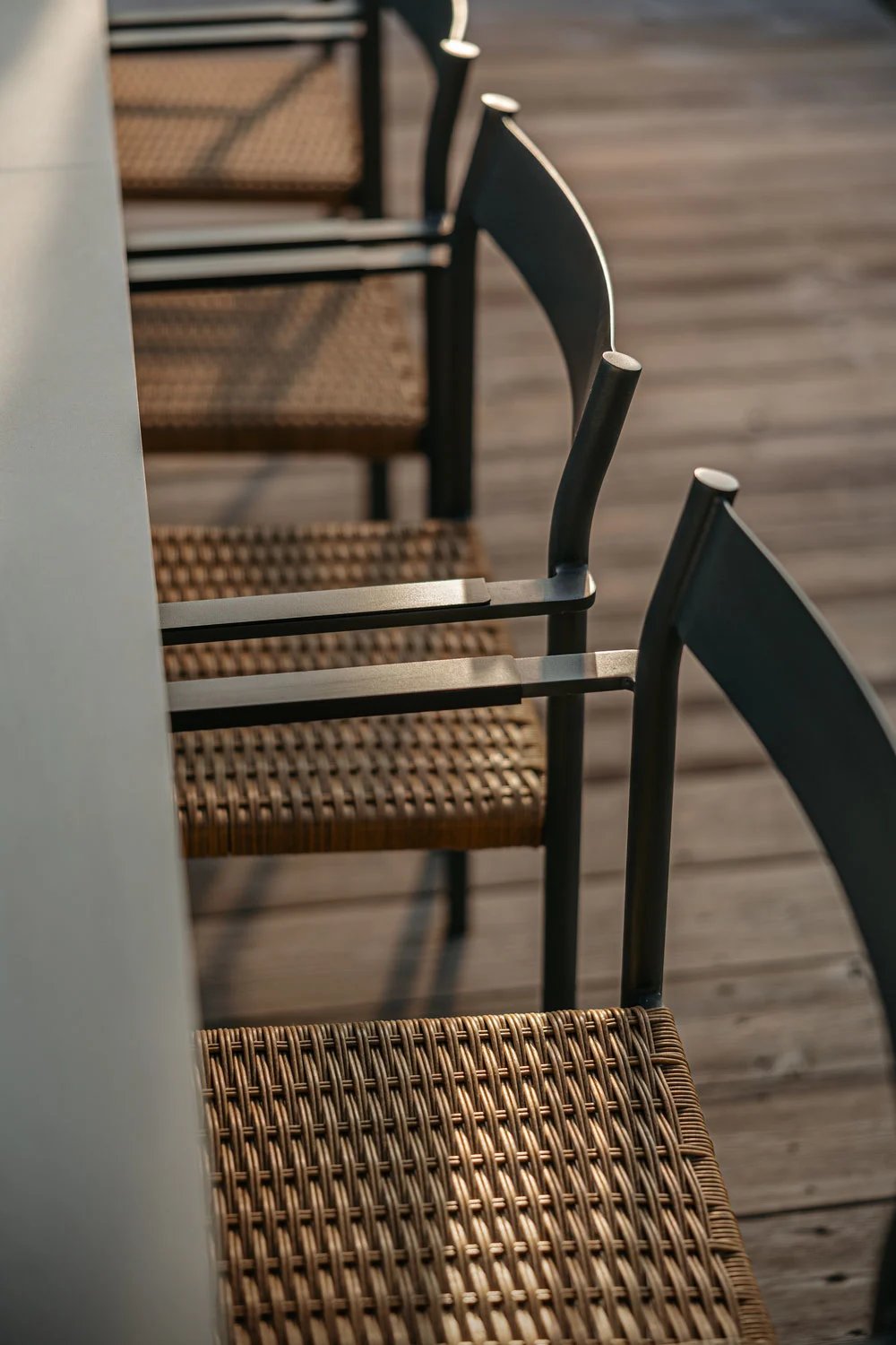Close-up of outdoor chairs with woven seats and dark metal frames on a wooden deck, bathed in warm sunlight.