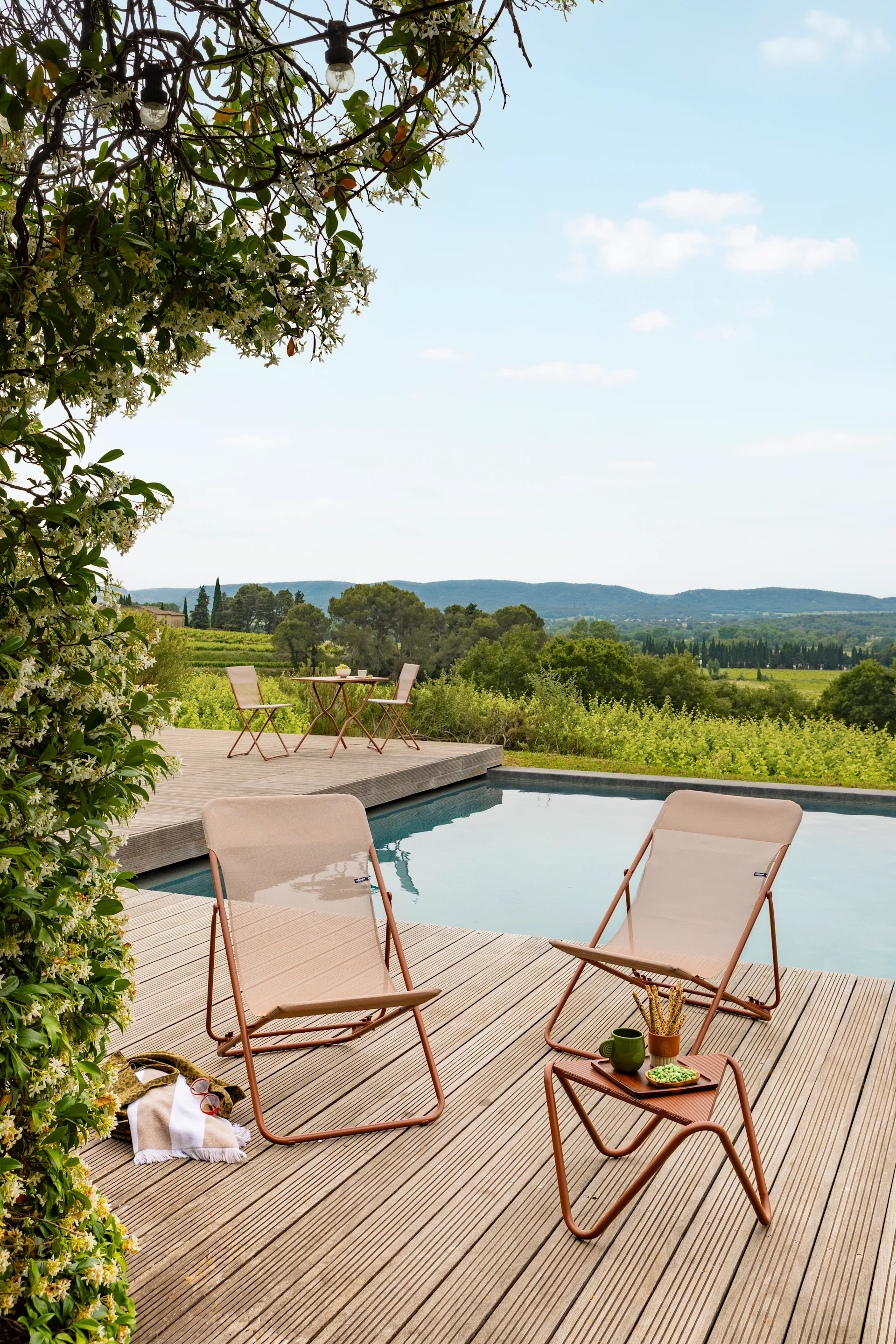 Two tan outdoor lounge chairs and a side table on a wooden deck by a pool, with flowering vines and a vineyard view.