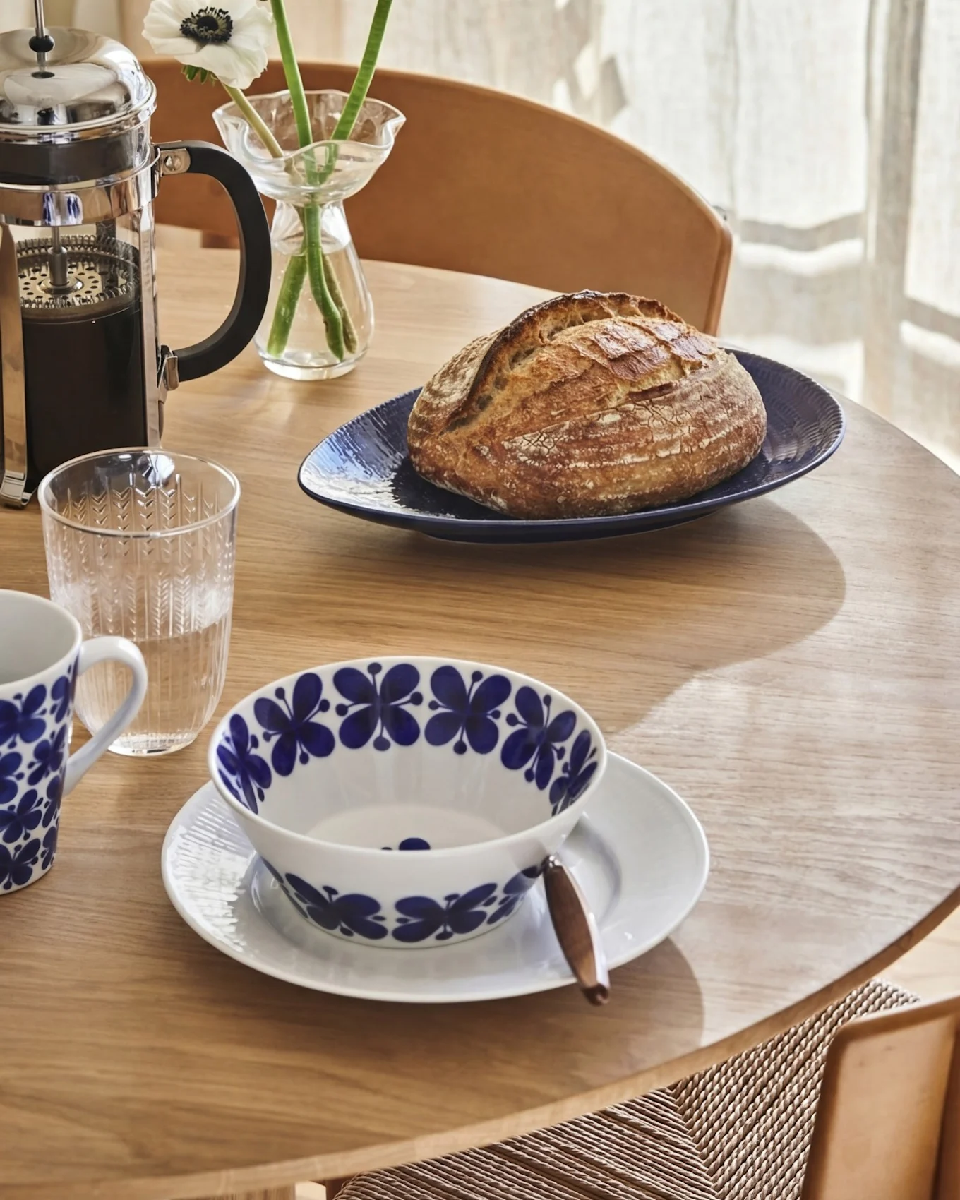 Breakfast table with French press, artisan bread, water glass, and patterned bowl and mug.