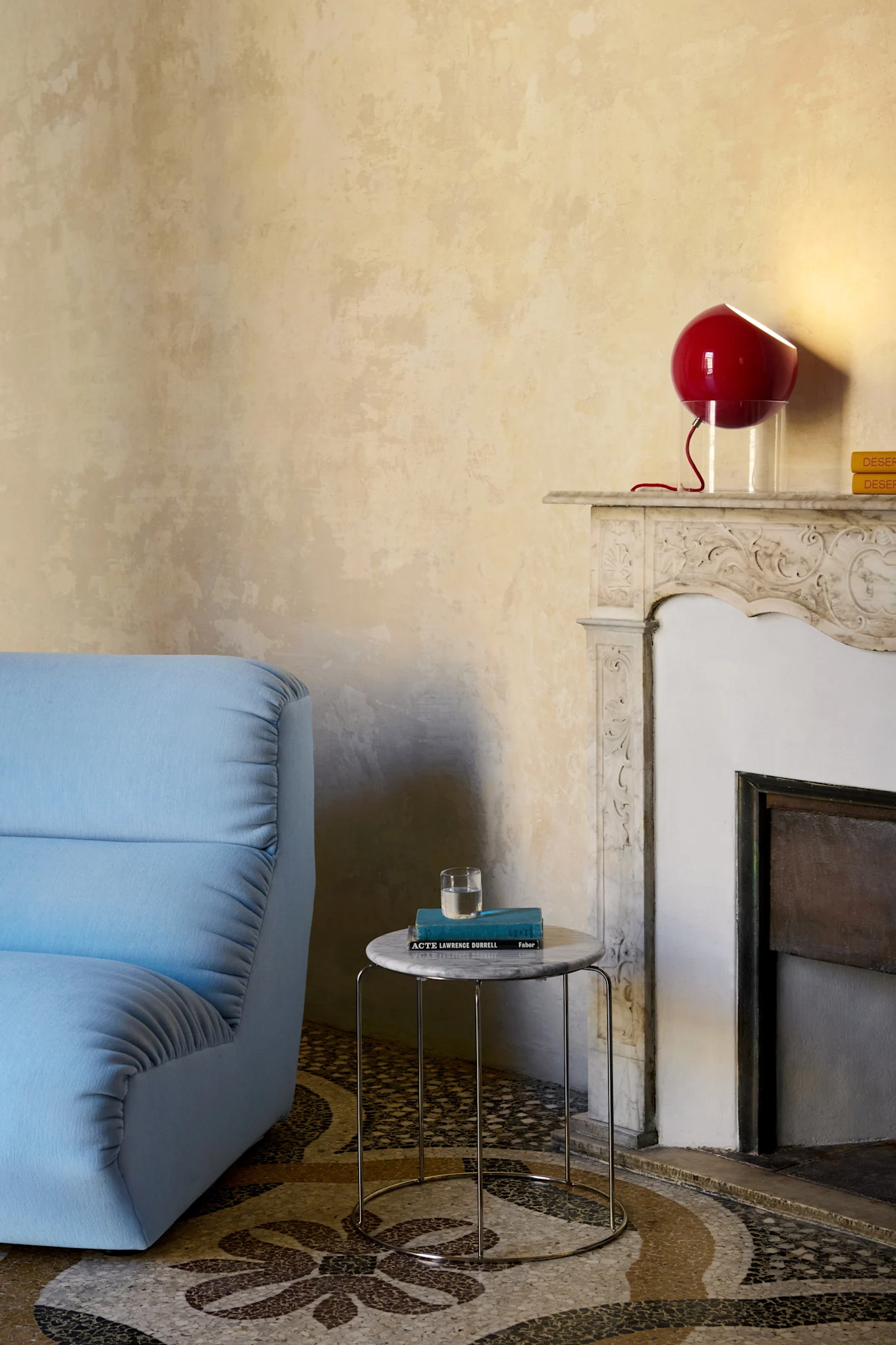 A blue sofa placed next to a fireplace, with a red lamp on top that adds a playful and unexpected expression and reflects the interior design trends of 2026.