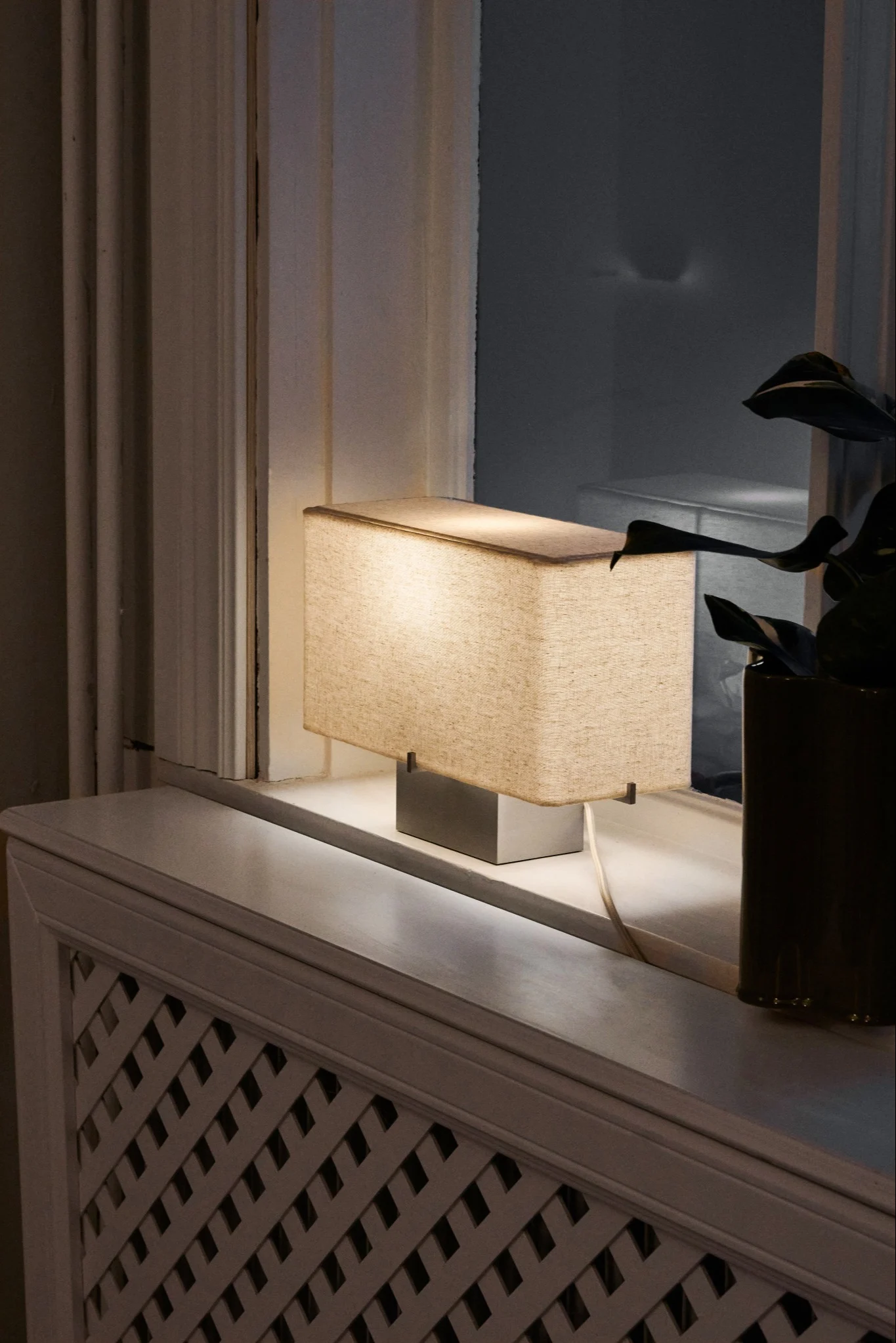 Rectangular table lamp with beige fabric shade and silver base on a white windowsill, next to a window and plant.