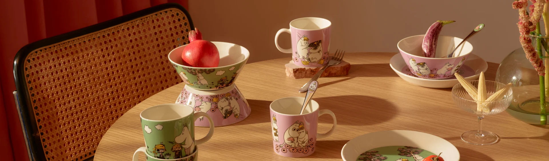 A wooden table set with Moomin-themed green and pink mugs, bowls, and plates, alongside a pomegranate, eggplant, and baby corn.