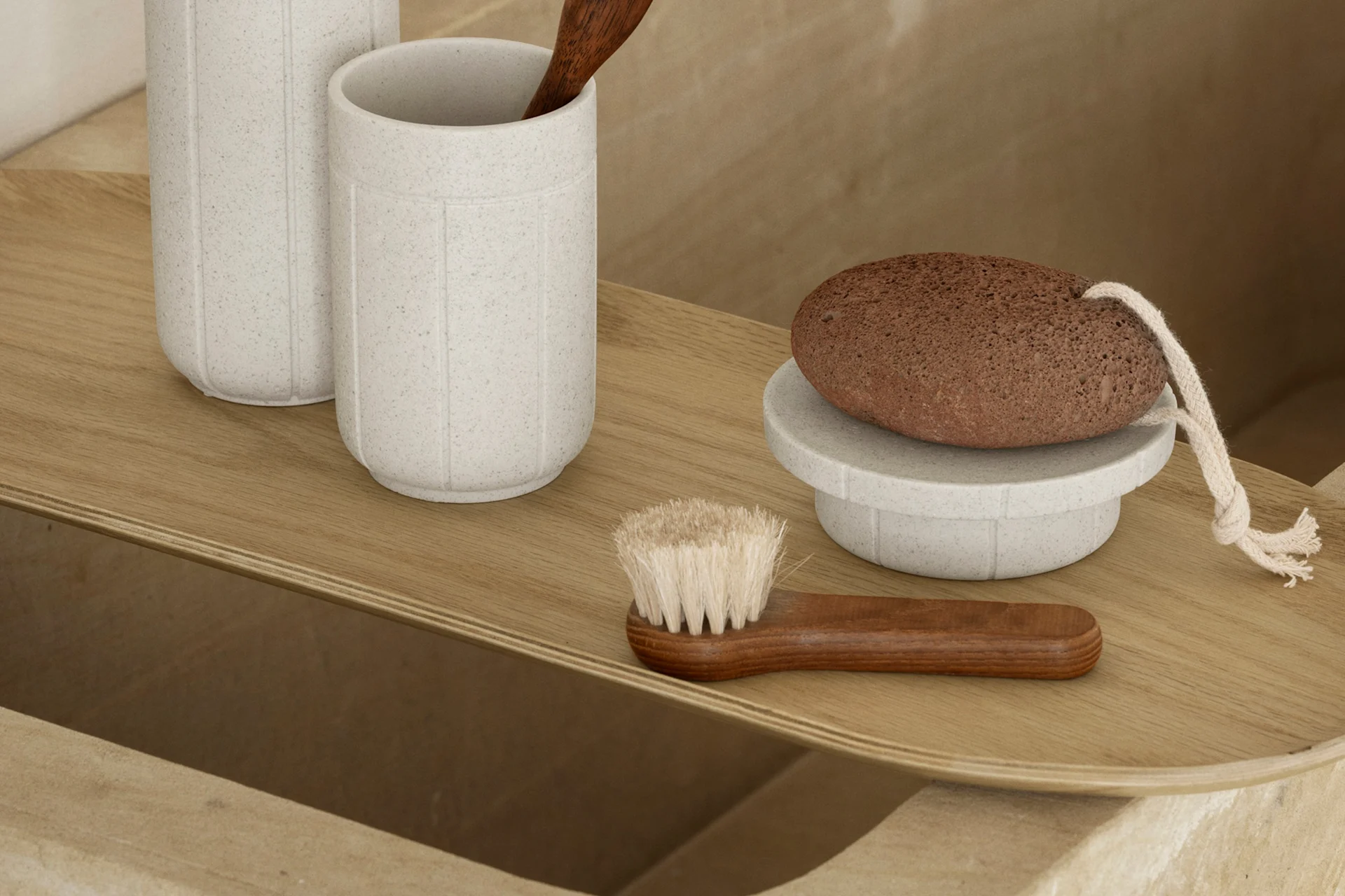 A light wooden tray holding bath accessories: two speckled grey cups, a wooden spoon, a brown pumice stone on a stand, and a wooden body brush.