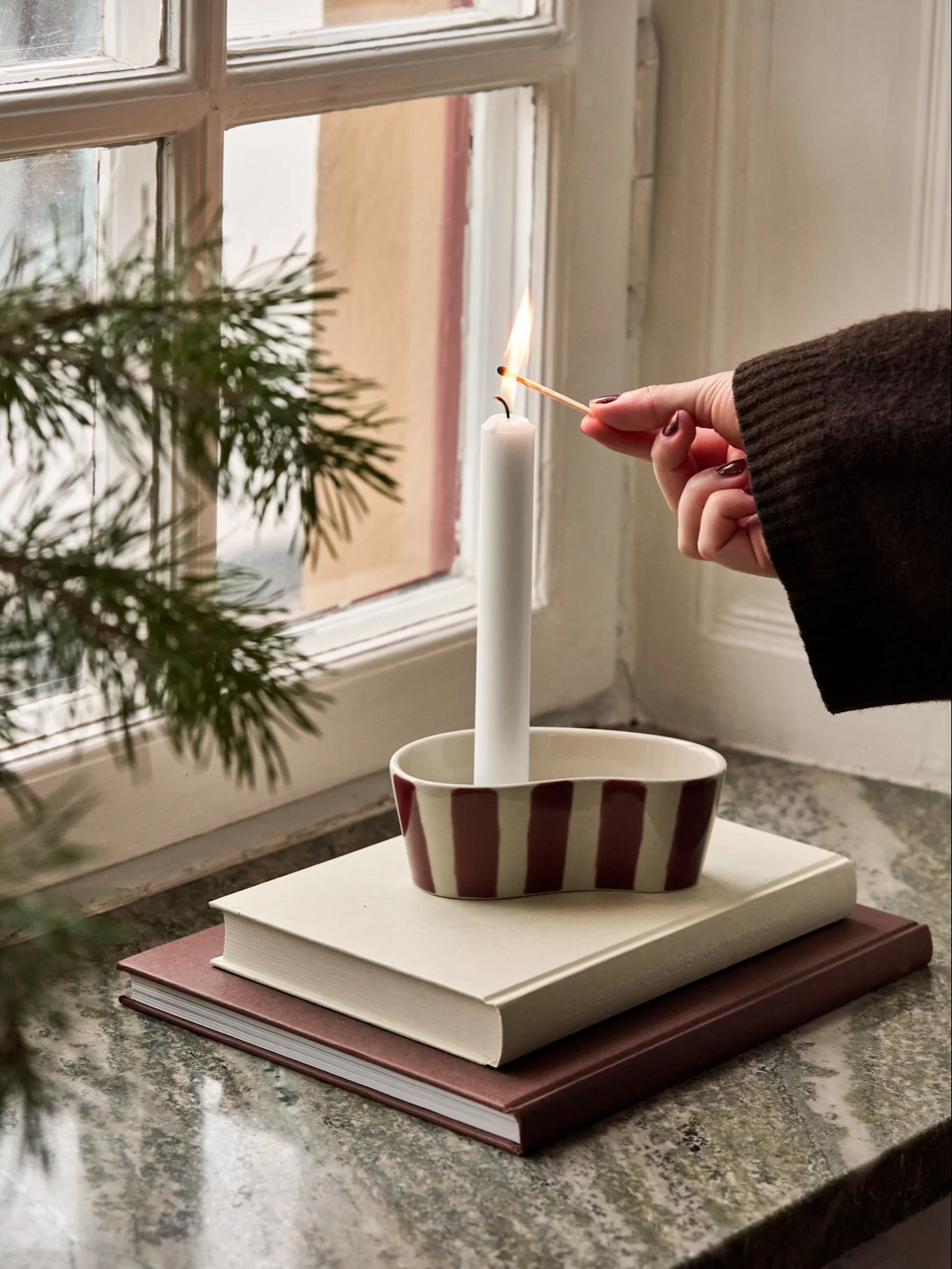 A hand lights a white candle in a striped holder on a stack of books by a window with pine branches.