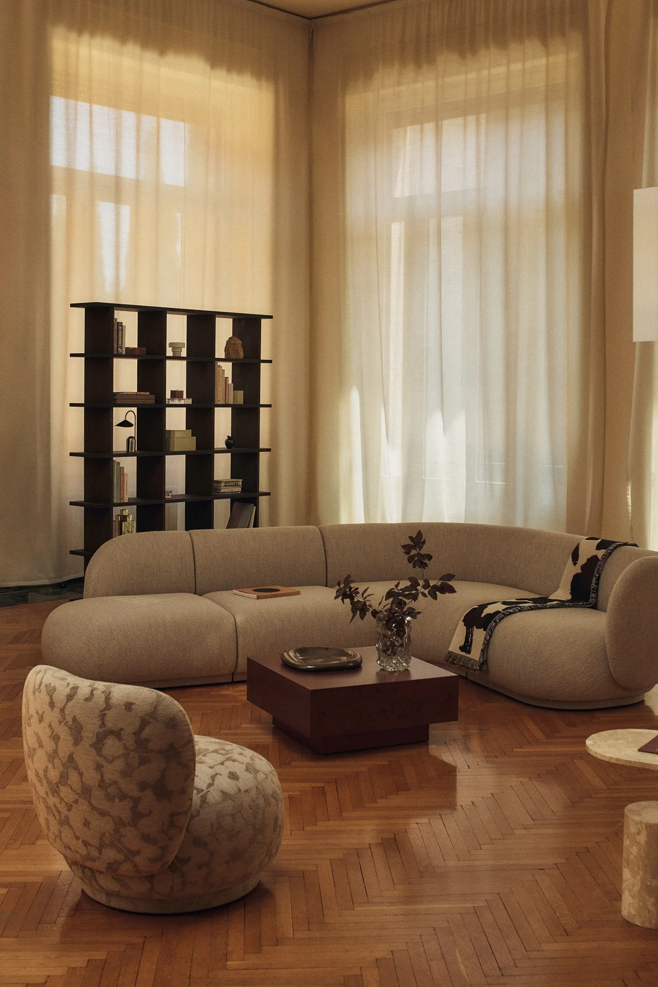 A living room with a curved beige modular sofa, a dark square coffee table, a dark open bookcase, and a patterned accent chair on a parquet floor.