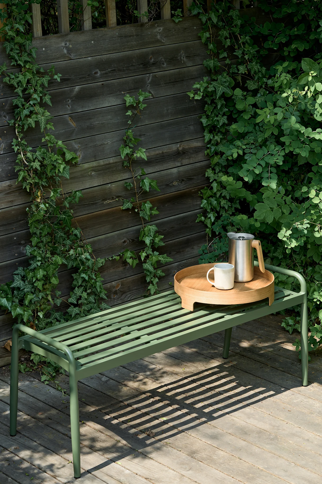Green slatted metal bench on a wooden deck with a wooden tray, white mug, and stainless steel carafe, next to an ivy-covered fence.