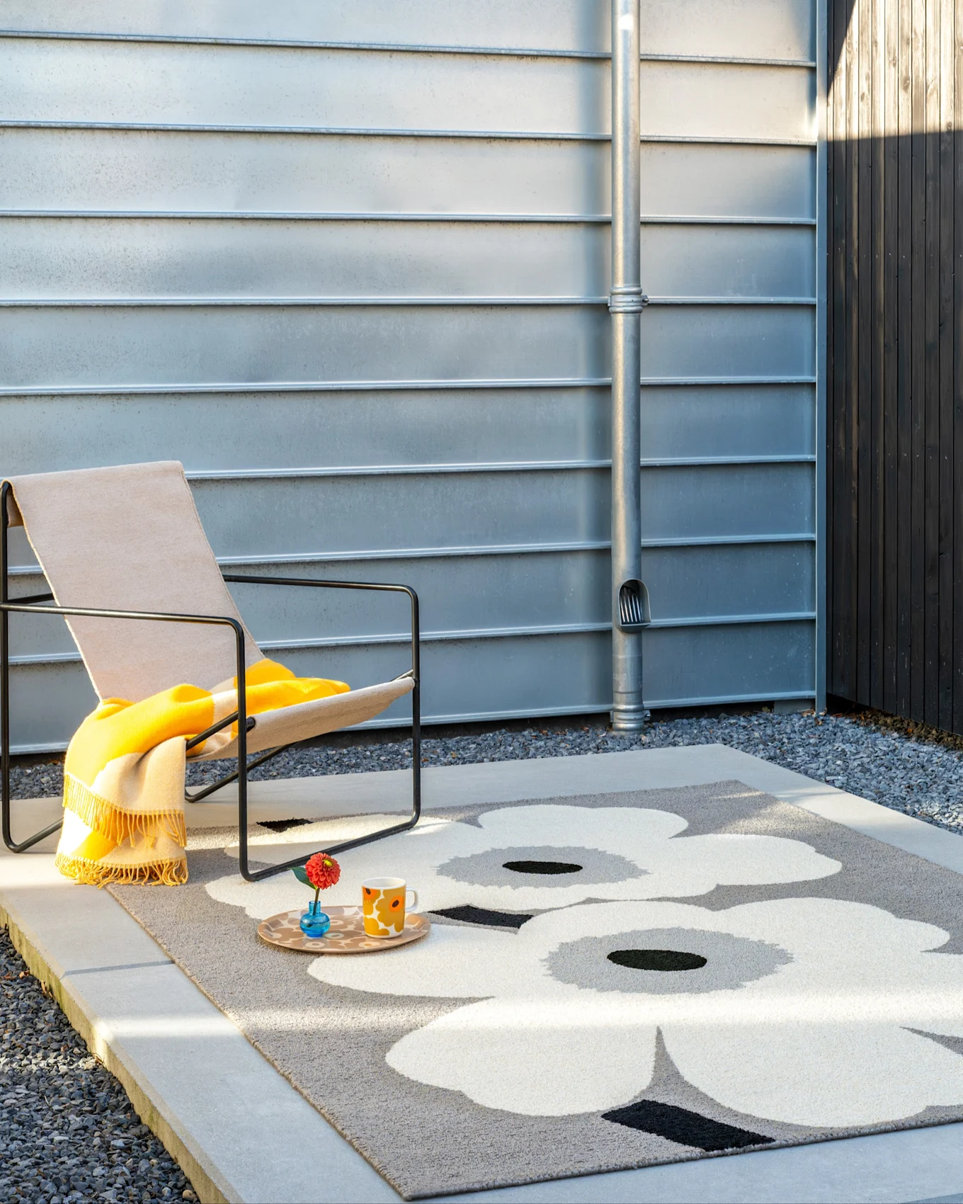 An outdoor scene with a dog walking on a grey rug with large white flowers, beside a chair with a yellow blanket and a metal wall.