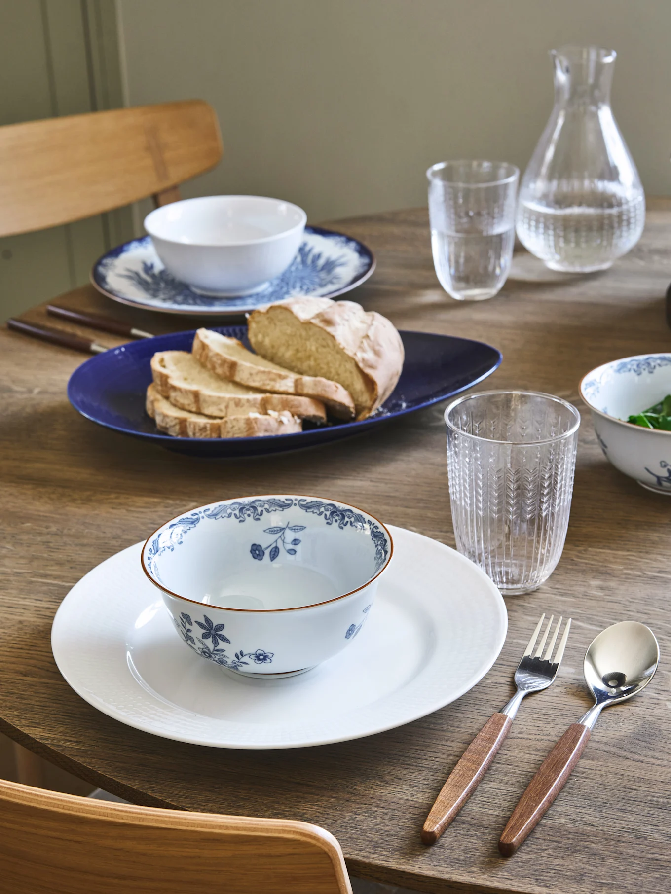 Dining table set with two place settings, blue and white floral bowls, bread on a blue plate, and wooden-handled cutlery.