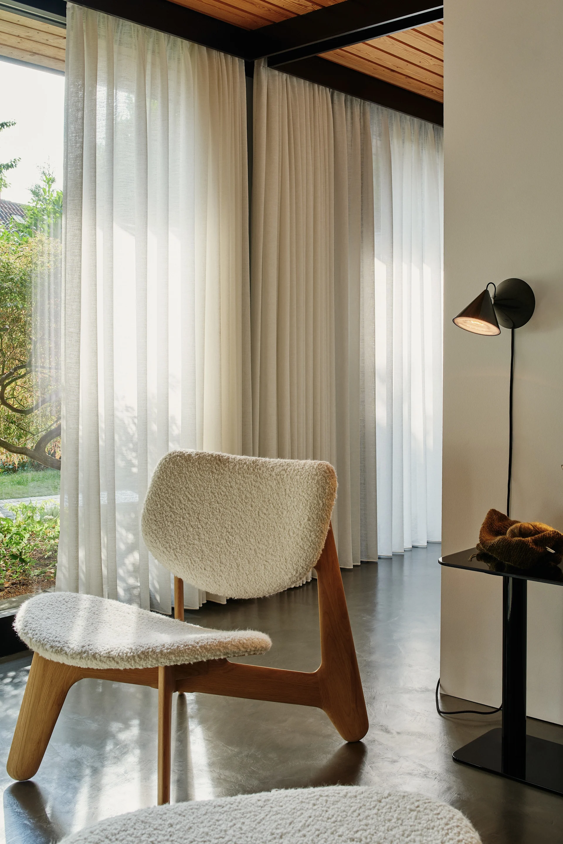 A white sheepskin armchair with wooden legs in a bright room with sheer curtains and garden view. A black wall lamp and side table.