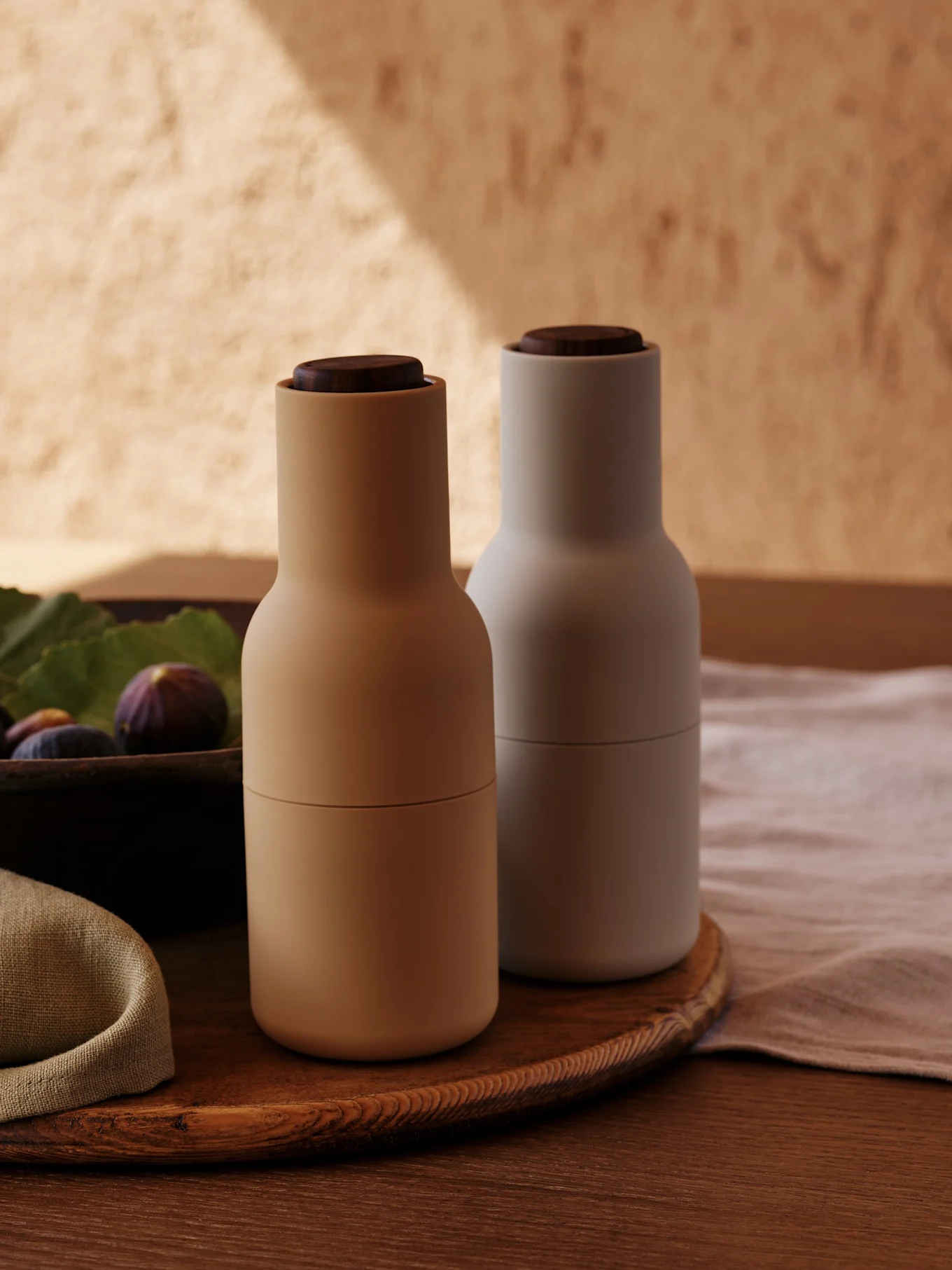 Two modern spice grinders with wooden tops, one beige and one light gray, on a round wooden tray with figs.