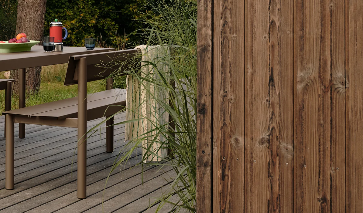 Brown metal outdoor table and bench on a wooden deck with fruit and drinks, next to tall grass and a wooden fence.
