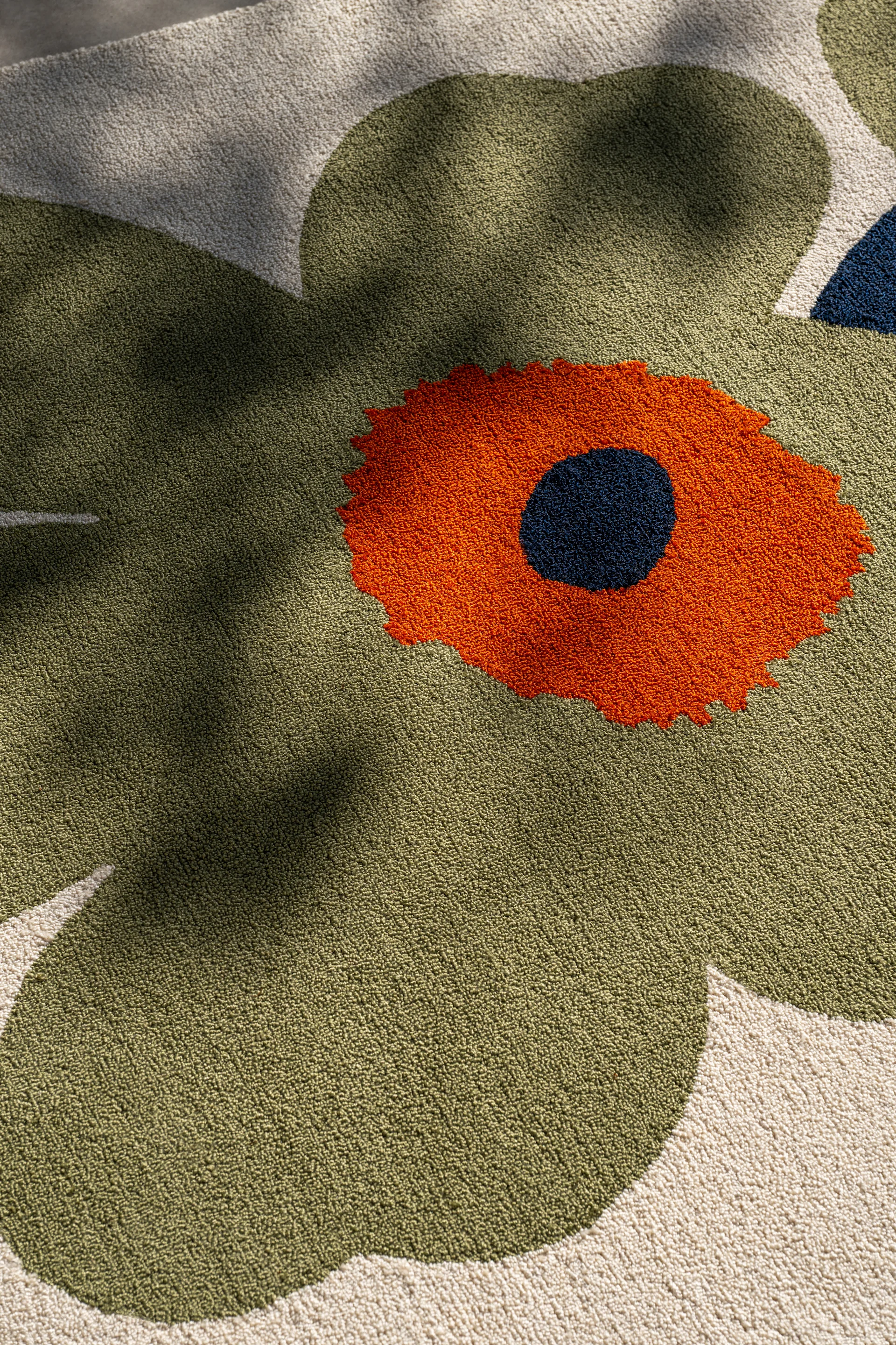 Close-up of a rug with a large, stylized flower design. Olive green petals, an orange center, and a dark blue dot are on a cream background.