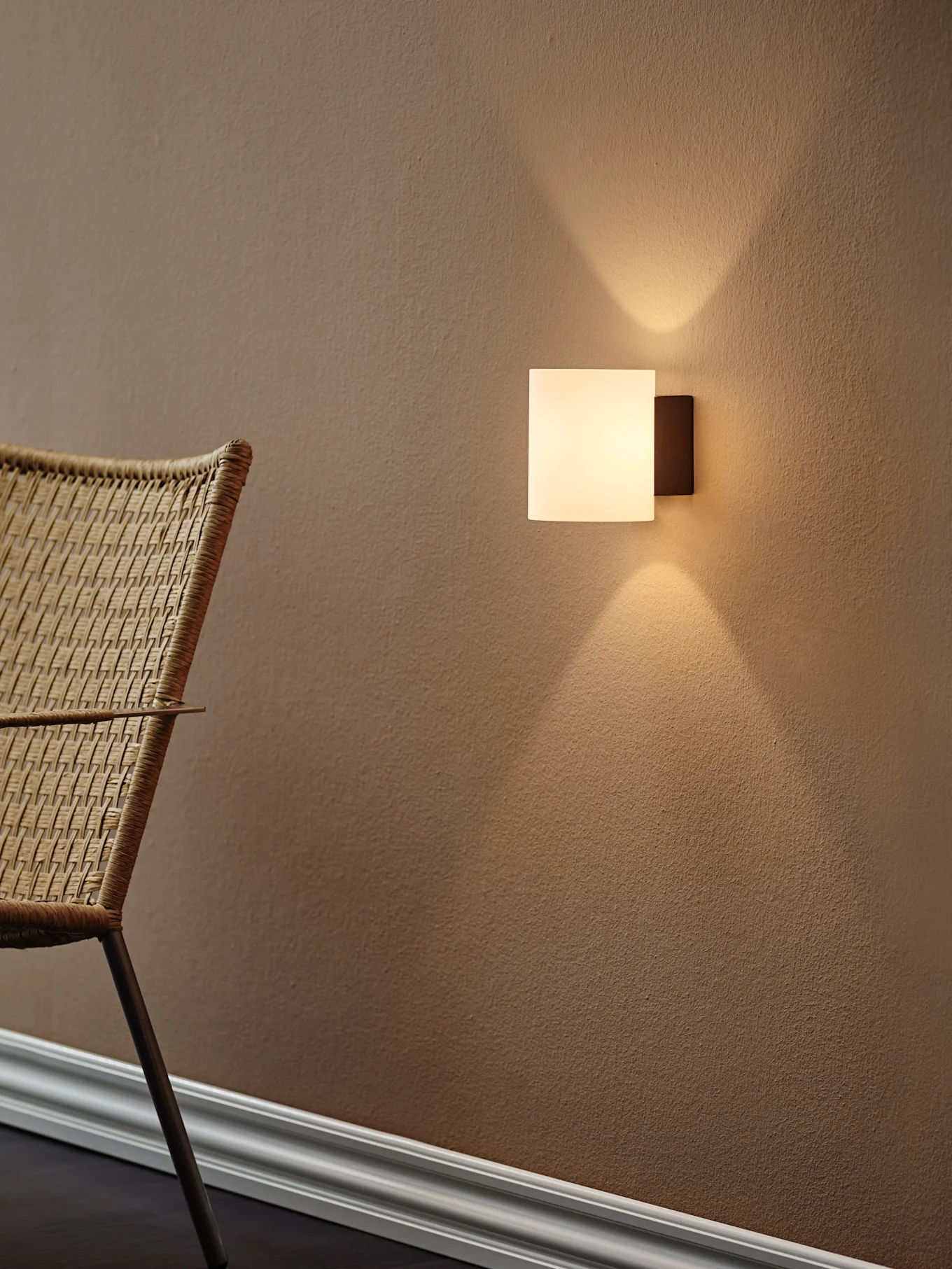 Modern square wall light casting warm light upwards and downwards on a textured brown wall, with a woven chair to the left.