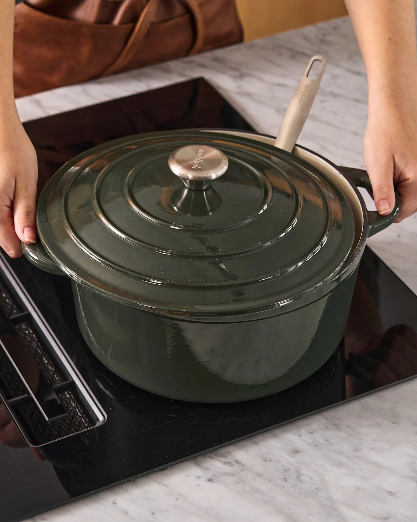 Person placing a dark green Dutch oven with a metal knob on a black induction cooktop.