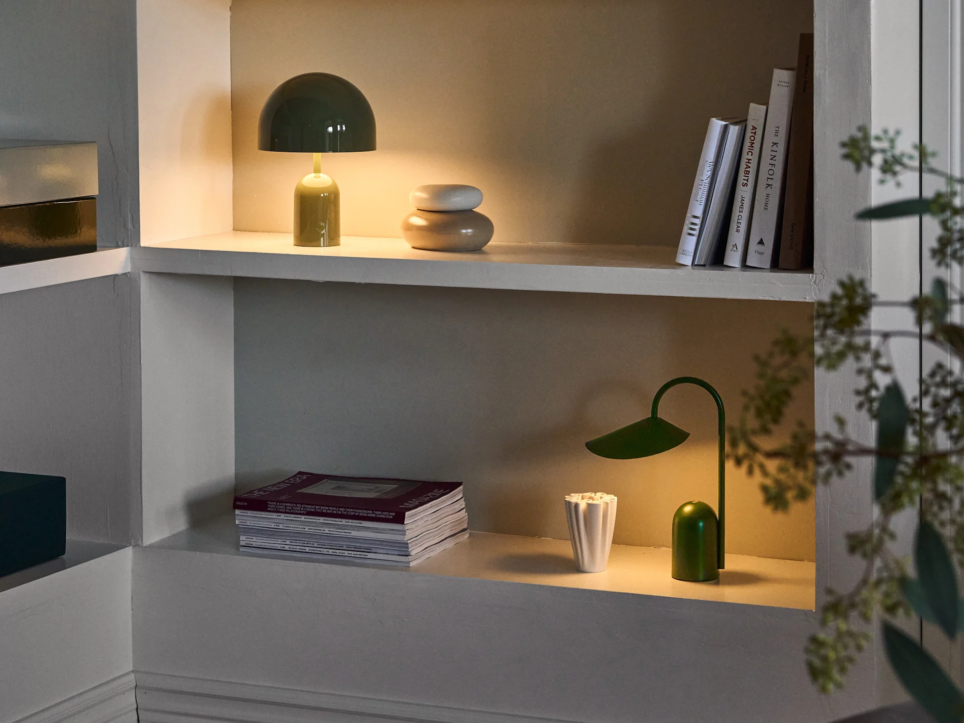 Illuminated shelves. Top: green mushroom lamp, stacked beige decor, and books. Bottom: magazines, white vase, and green leaf lamp.