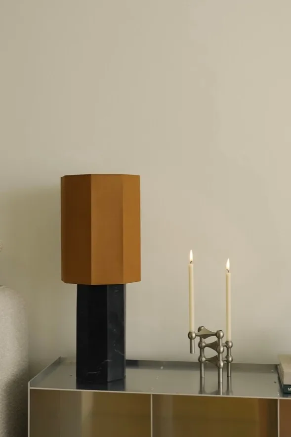 Modern table lamp with hexagonal brown shade and black marble base, next to a silver modular candle holder with two lit white candles.